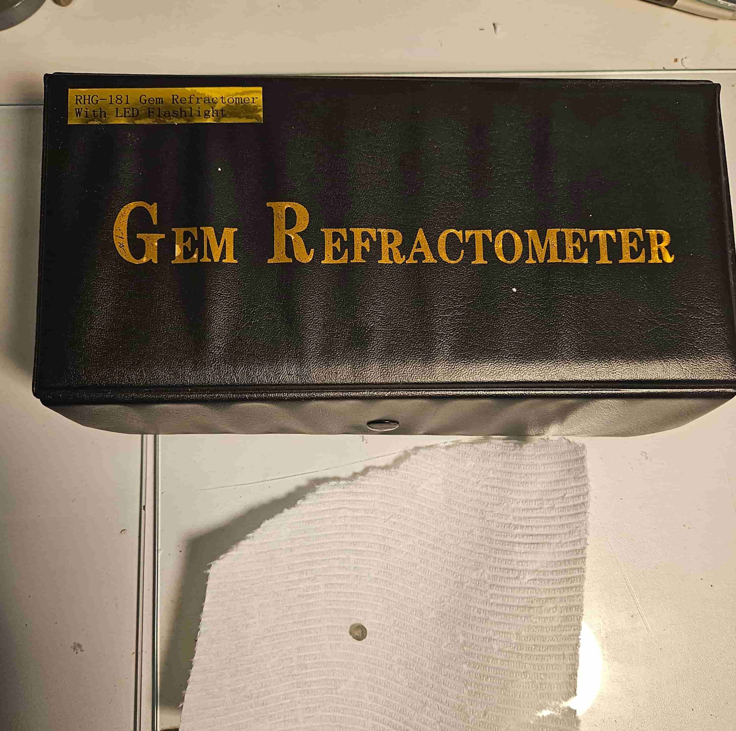 The image shows a black box with gold lettering that reads 'GEM REFRACTOMETER'. A torn piece of white paper towel with a sapphire on it is also visible beneath the box.