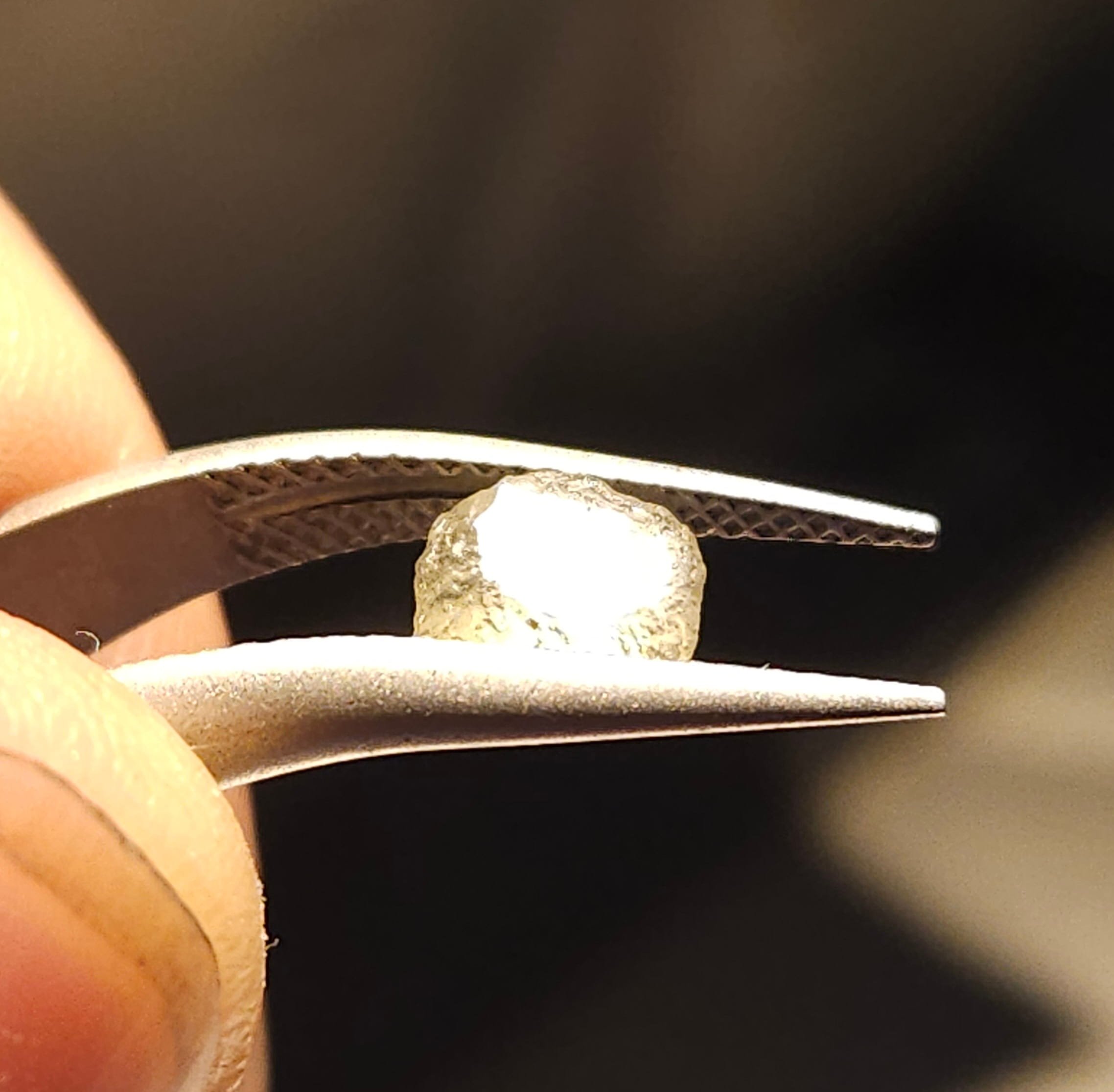 Close-up of a Sapphire held with tweezers, with part of a thumb visible in the foreground, and a dark background.