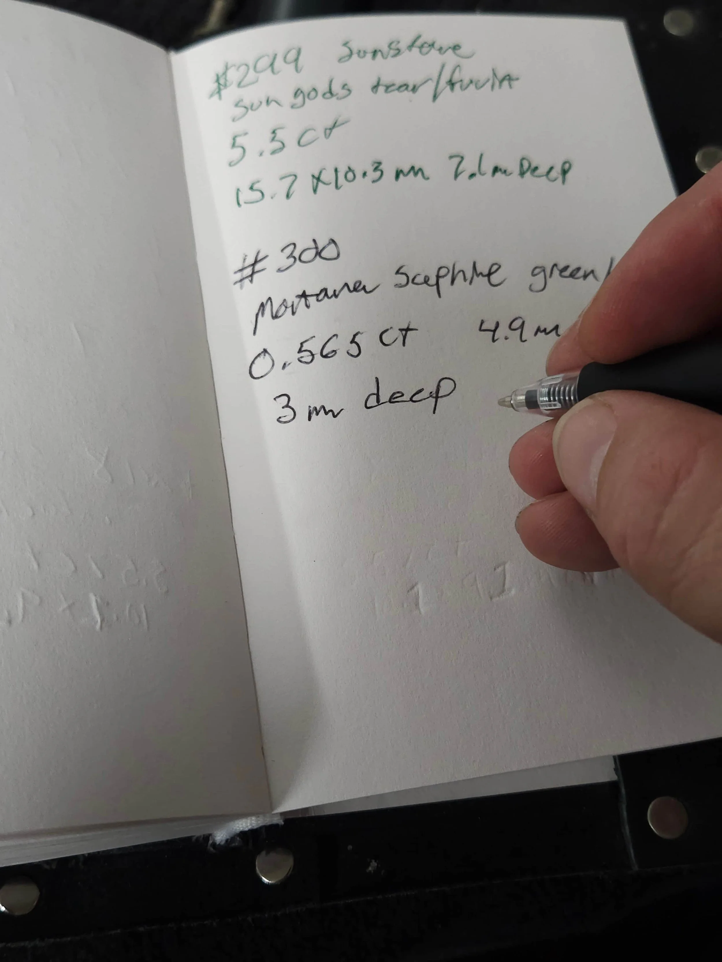 A person's hand holding a black pen and writing on a white notebook with handwritten notes in green and black ink. The notes include information about minerals and gemstones.