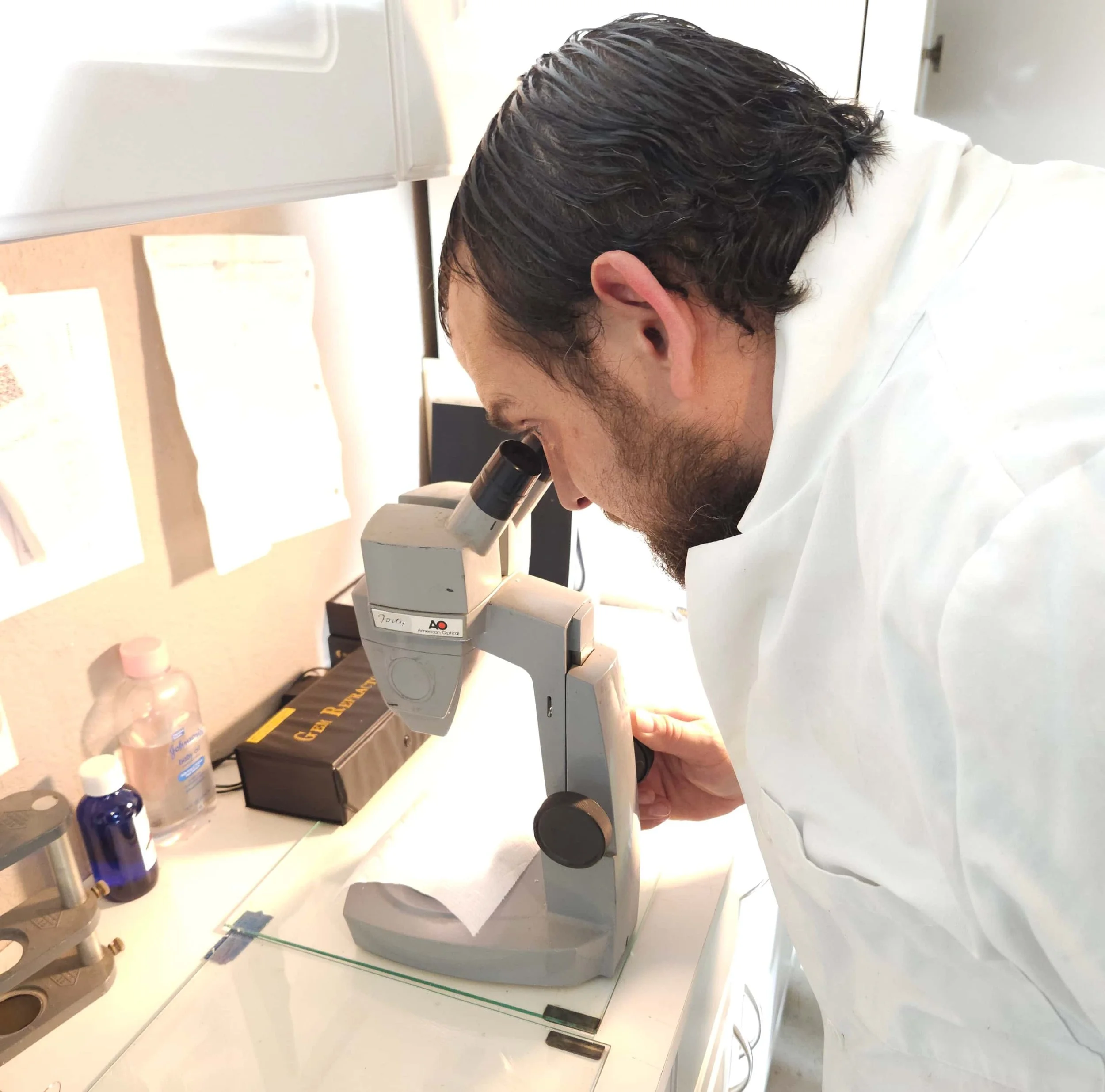Ian Perry from The Lapidary Office looking through a microscope on a white lab bench, surrounded by lab supplies including bottles and equipment.