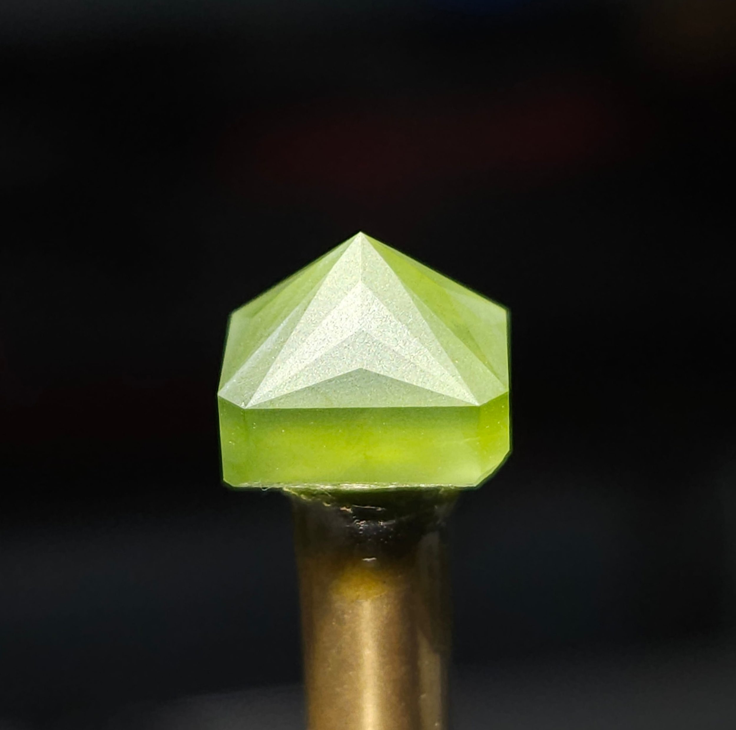 Close up of a peridot pavilion being cut at 500 grit on a brass dop attatched by green wax with a dark blurred background.