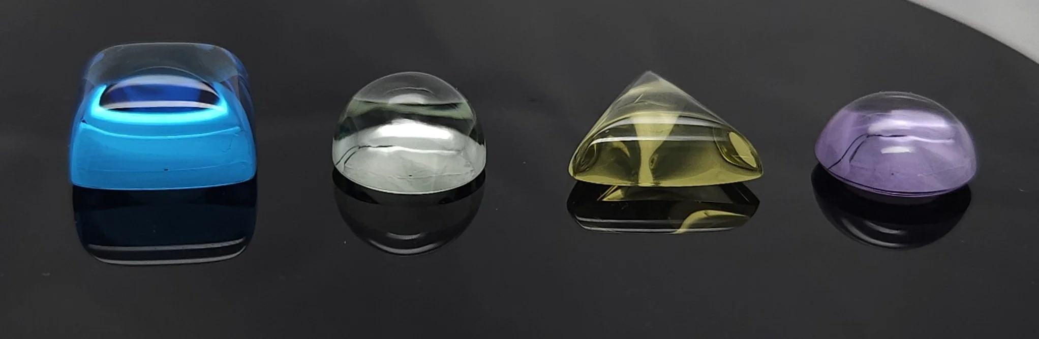 Four colorful Quartz cabochon stones on a reflective black surface, arranged in a row. The stones are blue, green, yellow, and purple.