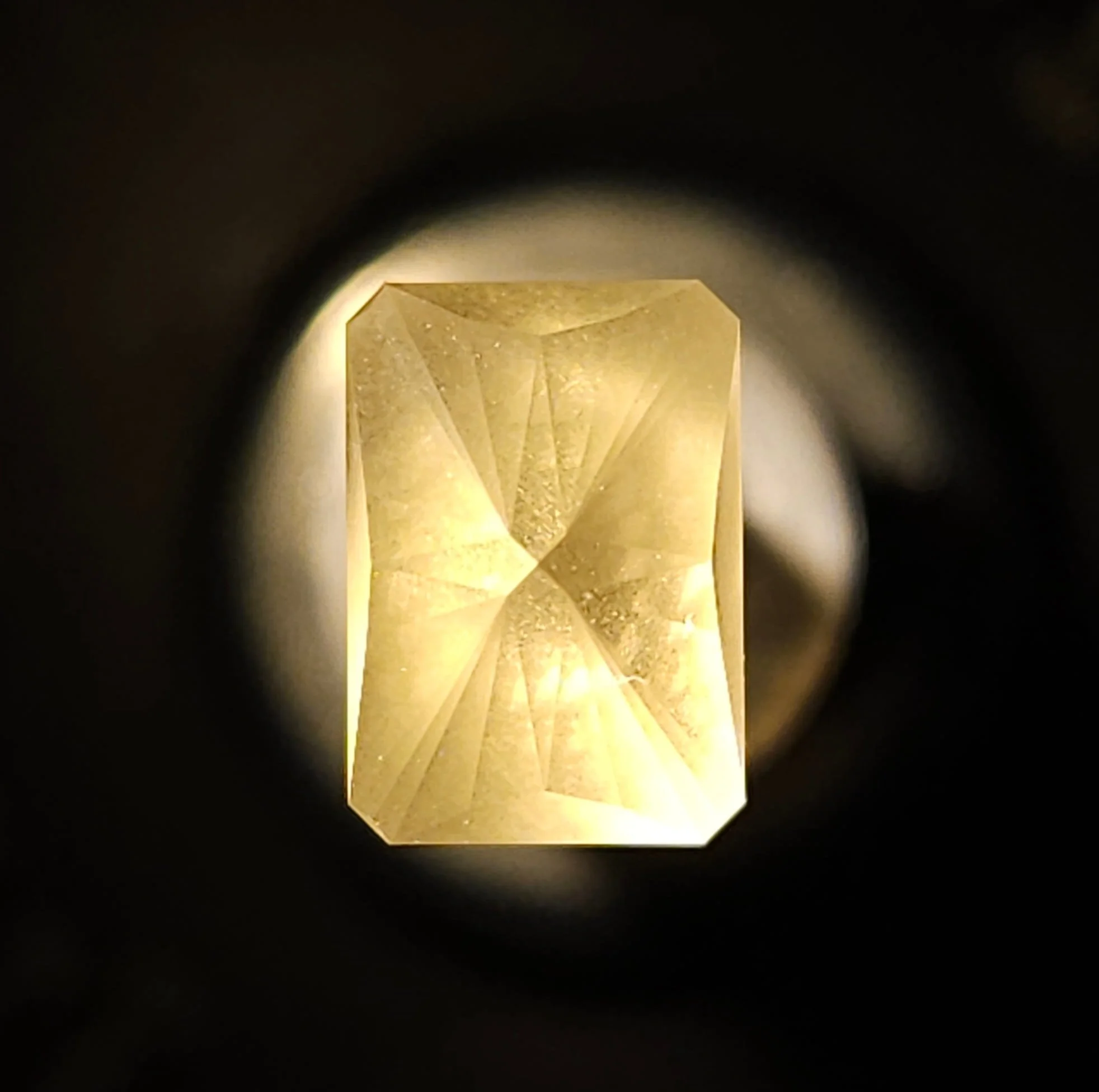 A gold-colored gemstone with a geometric cut, viewed through a circular aperture, with light illuminating the gemstone from below.