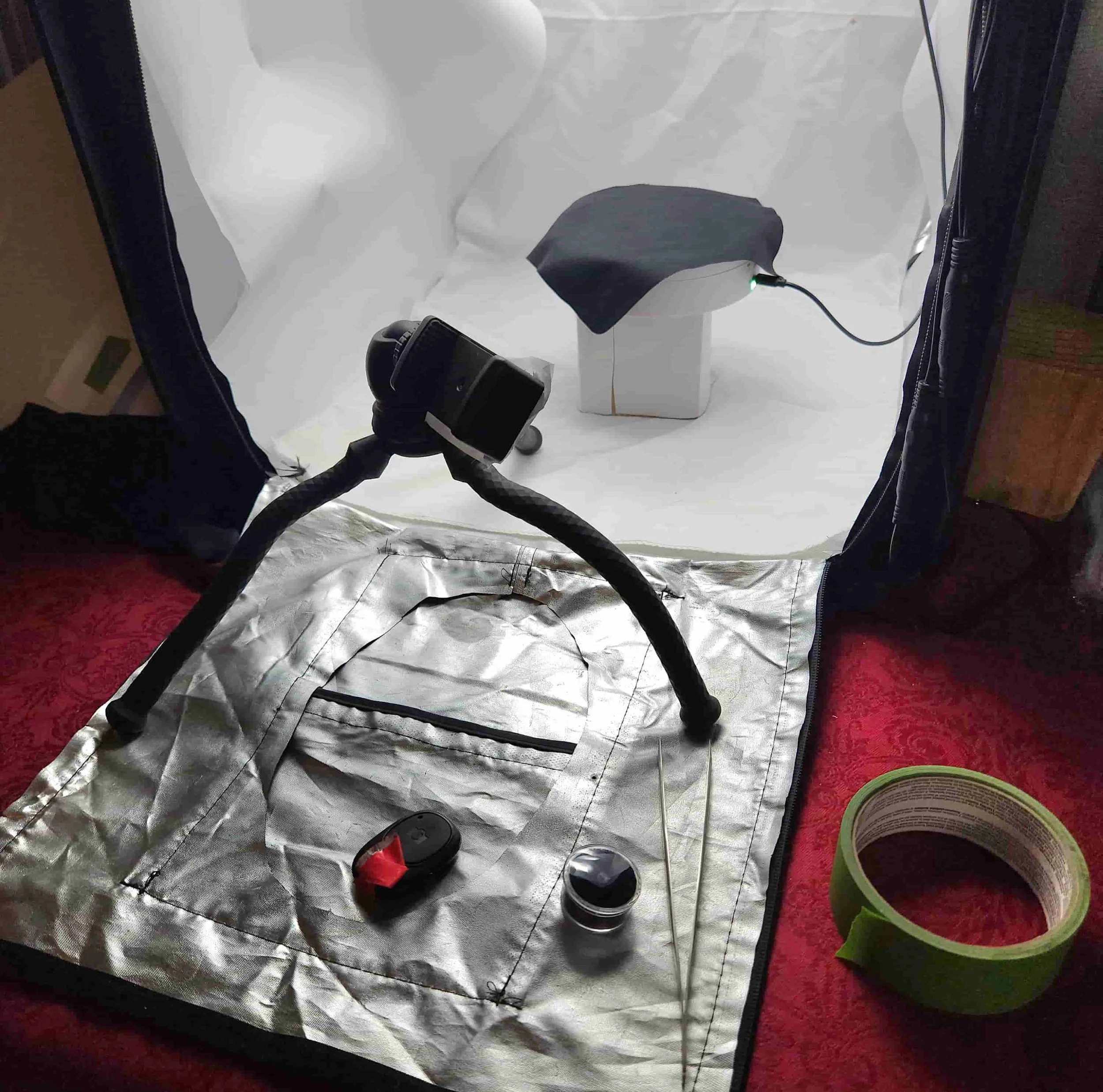 Inside a reflective photography tent, a small electronic device with a black cover and a green indicator light is connected to a power source through a cable. The tent has a crumpled white fabric background, with some tools and equipment on a red carpeted floor, including a black cylindrical object and a small container with purple contents. The scene appears to be set up for product photography or technical work.