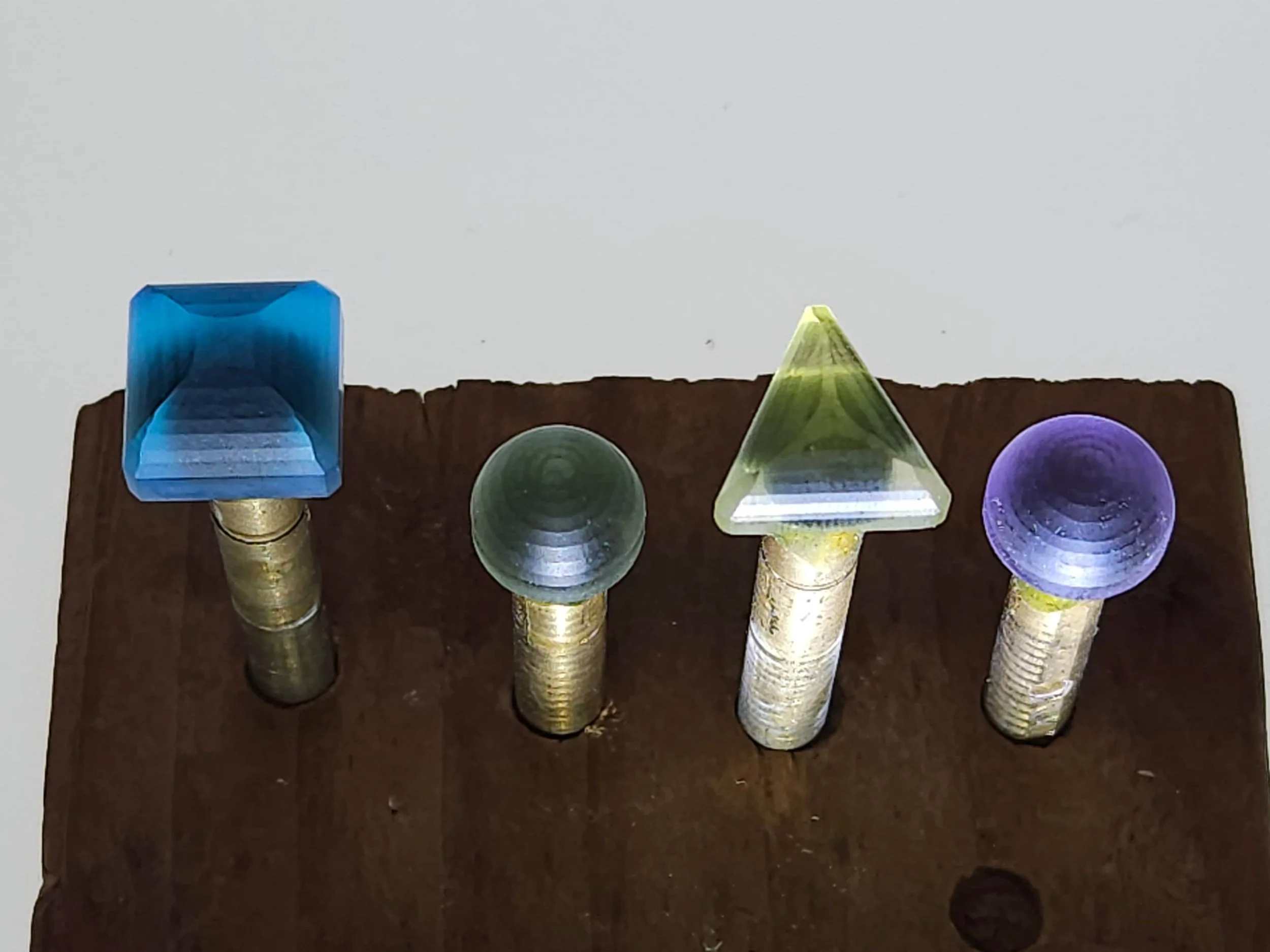 A display of seven small, colorful quartz cabochons attached to brass dops on a dark wooden surface, including a blue cube, a green sphere, a yellow cone, and two purple spheres.