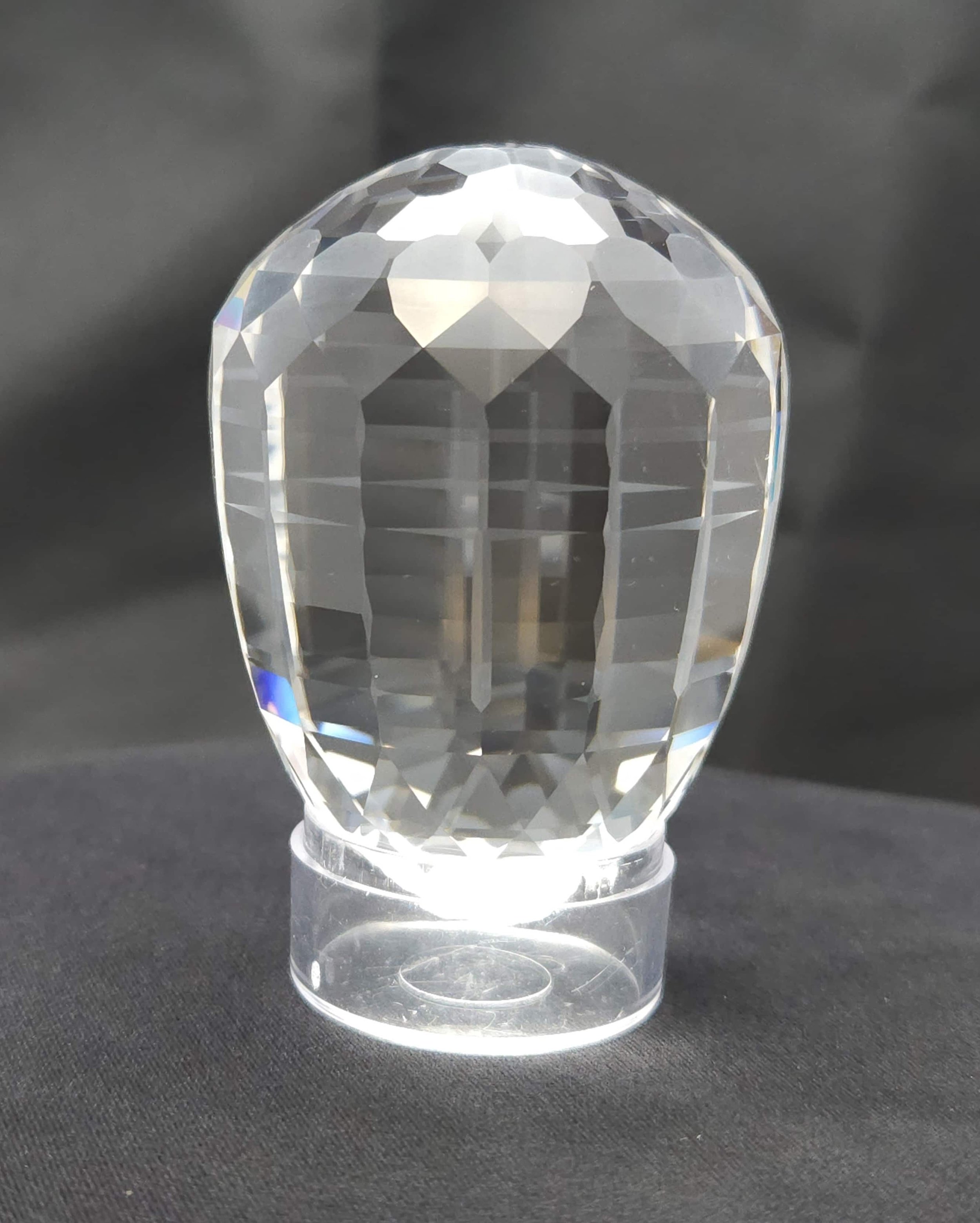 A clear faceted glass or crystal trophy or award with a rounded top, standing on a wide, flat base, placed on a black surface with a dark gray background.