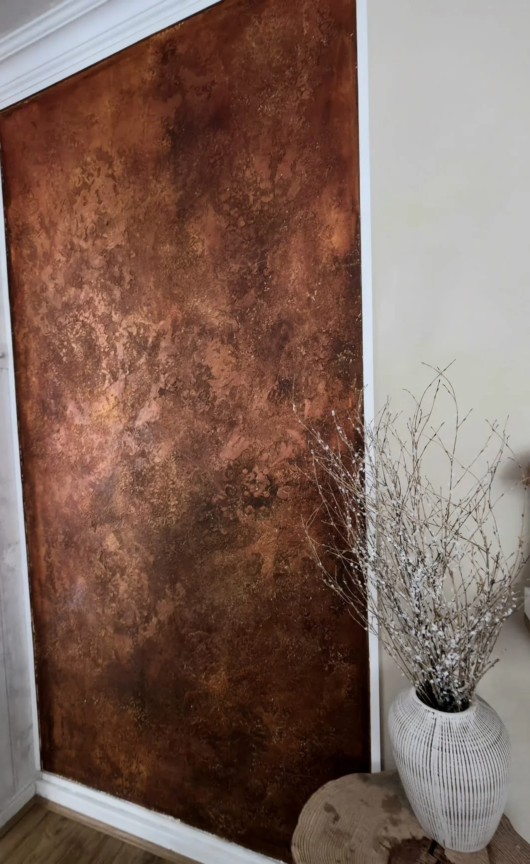 A brown and rust-colored textured wall panel next to a beige wall with a white vase holding dried branches.
