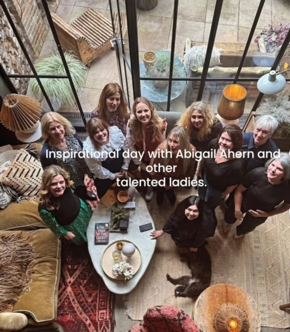 A group of women are gathered inside Abigail Ahern's living room, looking up at the camera while standing around a coffee table with books, candles, and a vase of flowers. The room has large windows with a view of an outdoor patio decorated with plan