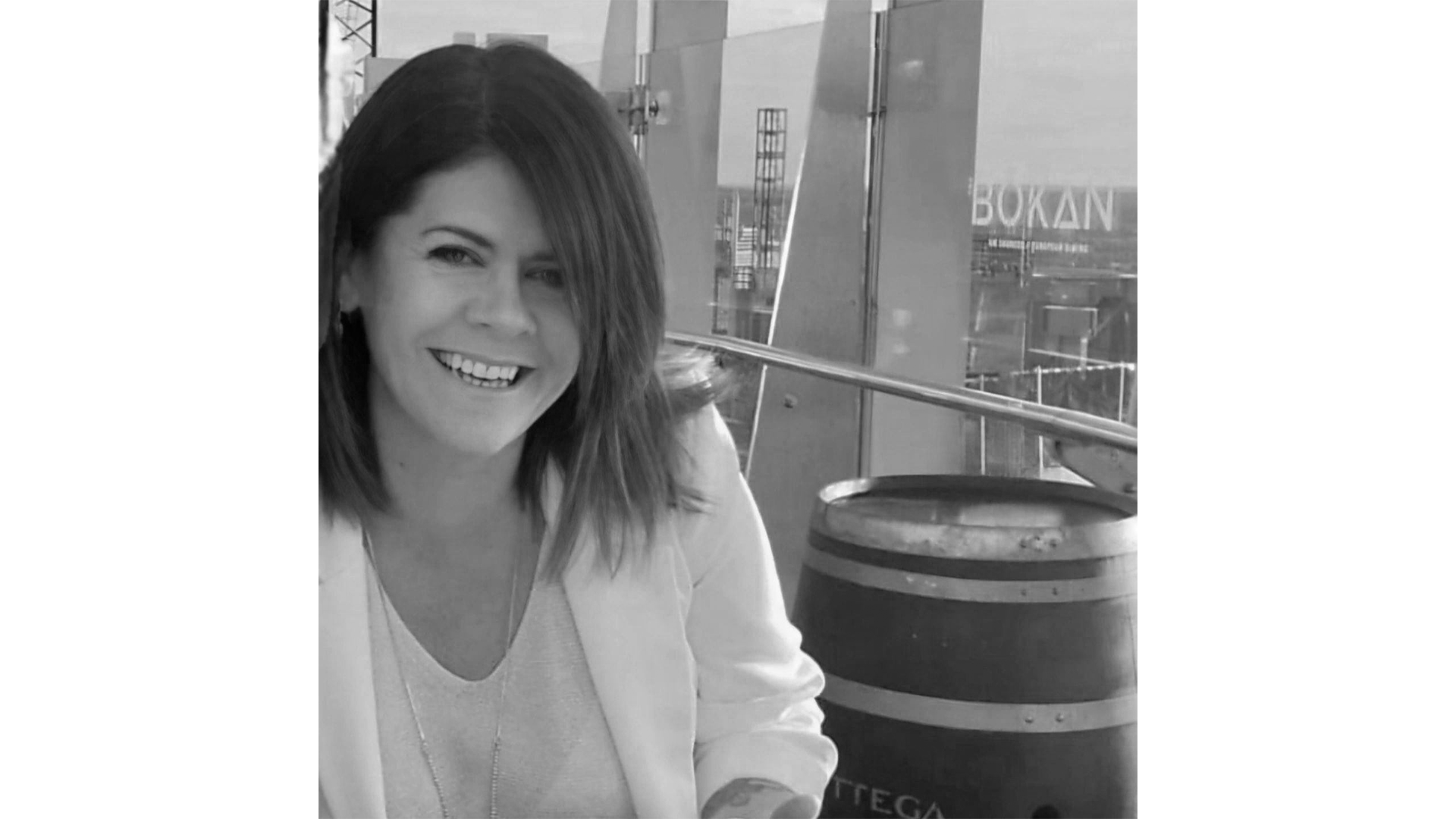 A woman with shoulder-length dark hair smiling, sitting near a beverage barrel, with a modern open-air setting in the background.