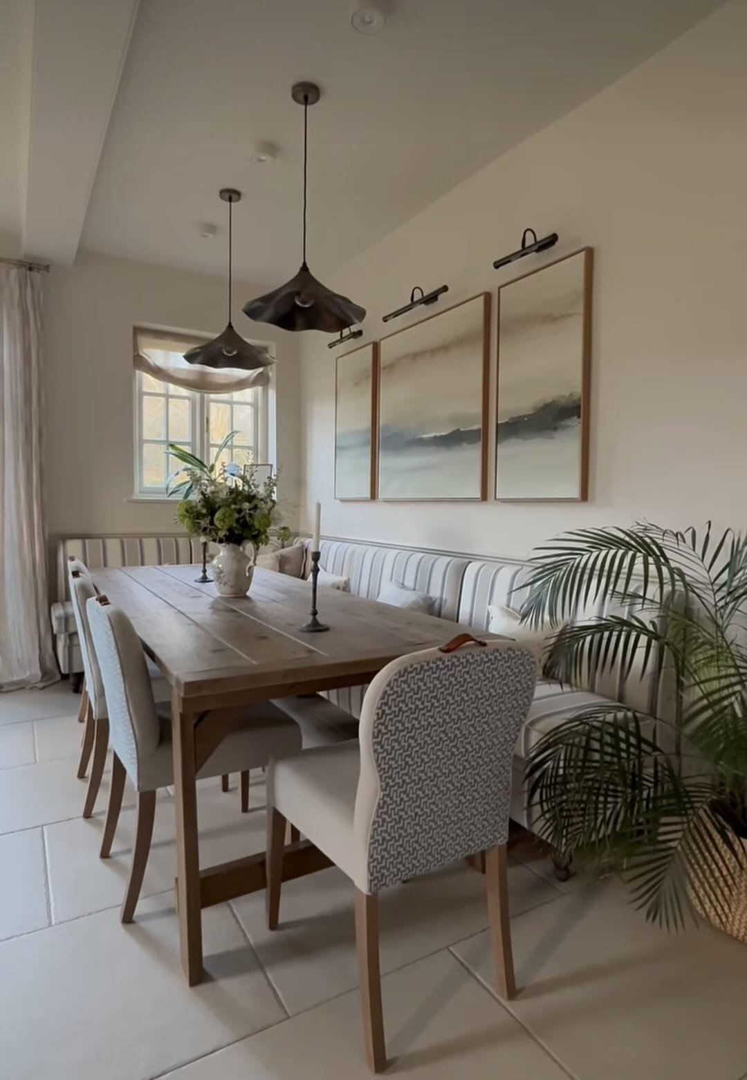 Dining room with a wooden table, floral centerpiece, two candlesticks, beige upholstered chairs, a striped bench, wall art, pendant lights, a window, and a potted plant.