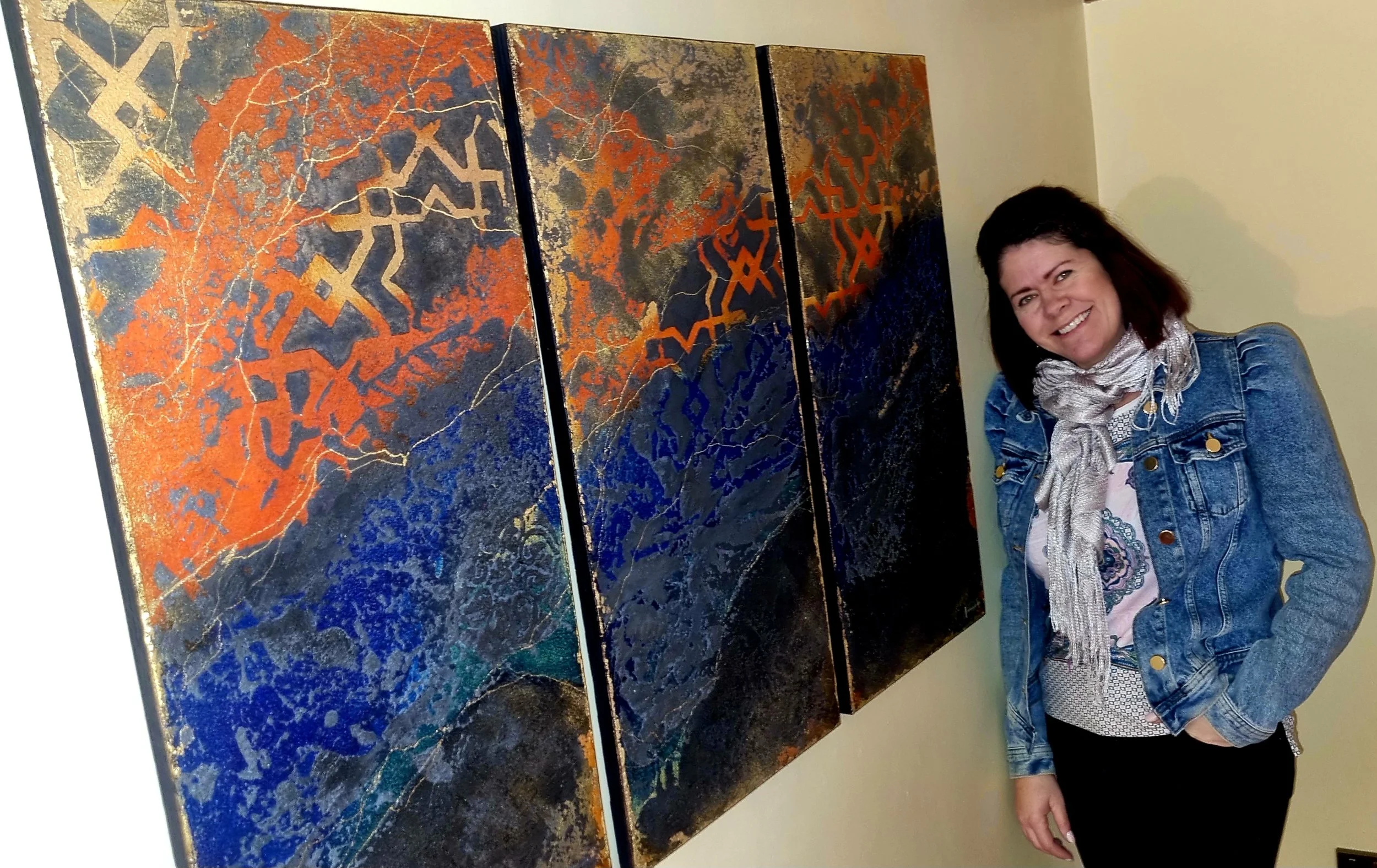 A woman smiling at the camera standing beside three abstract paintings with orange, blue, gold, and dark colors on the wall.