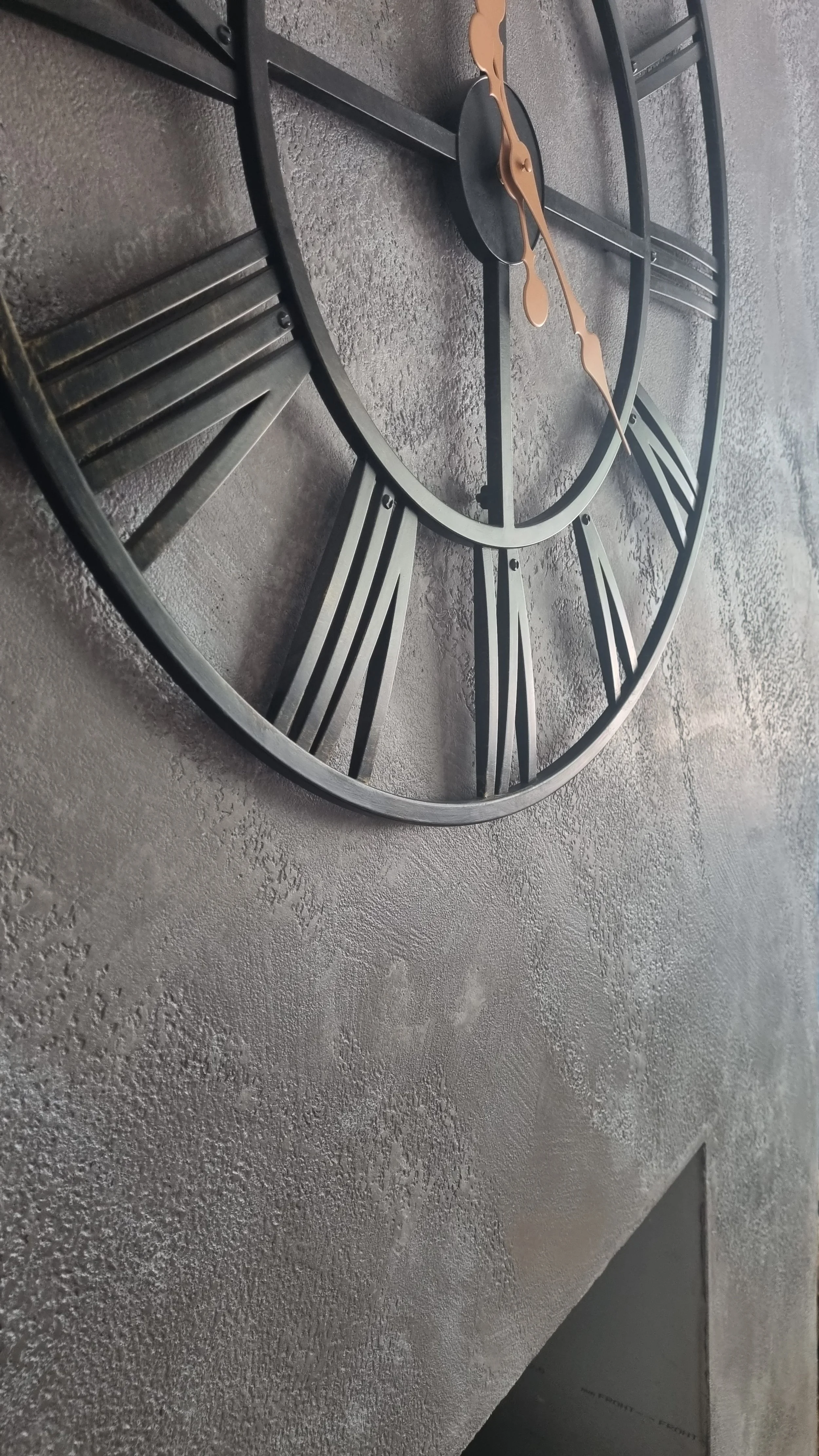 Close-up of a wall clock with a black metal frame and Roman numerals, placed on a textured Venetian plaster wall.