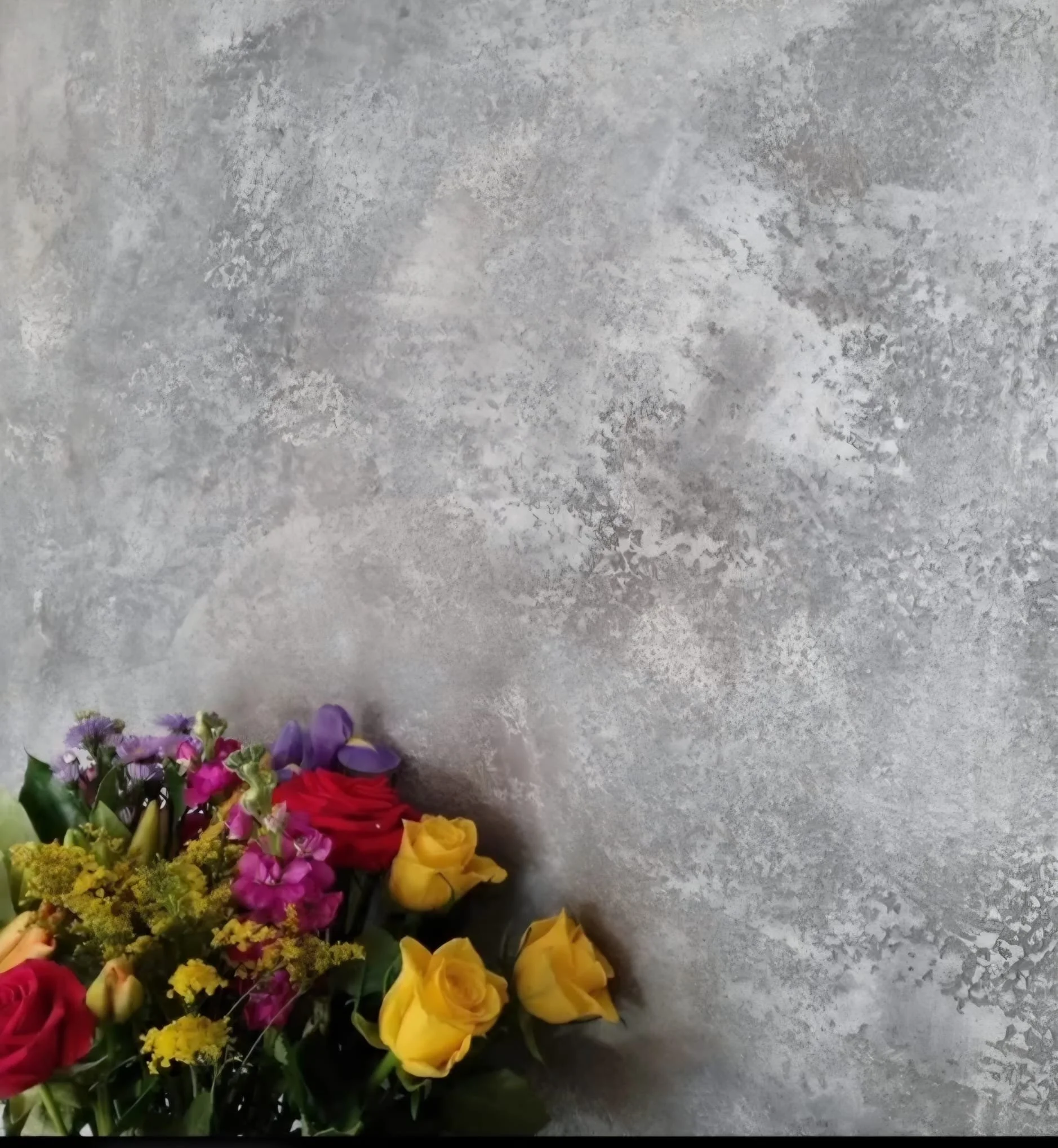 A mixed bouquet of flowers lying on a textured gray surface, including yellow roses, red roses, purple flowers, and other colorful blossoms.