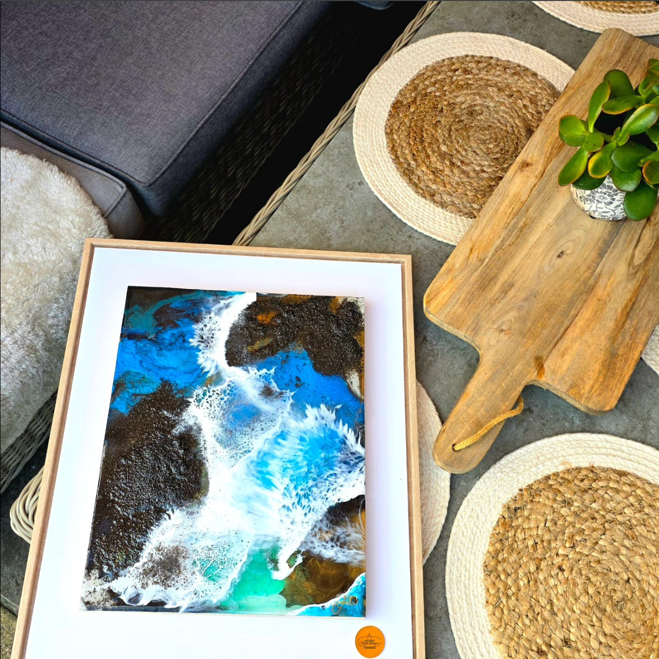A framed ocean shoreline painting, a wooden cutting board, a potted succulent plant, a woven straw placemat, and part of a gray sofa and a beige shaggy rug visible in the scene.
