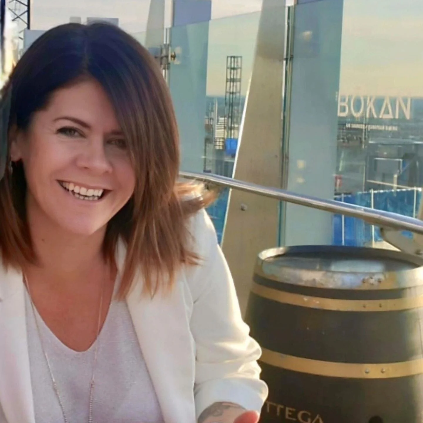 A woman with shoulder-length brown hair smiling, sitting outdoors near a glass barrier, with a cityscape in the background.