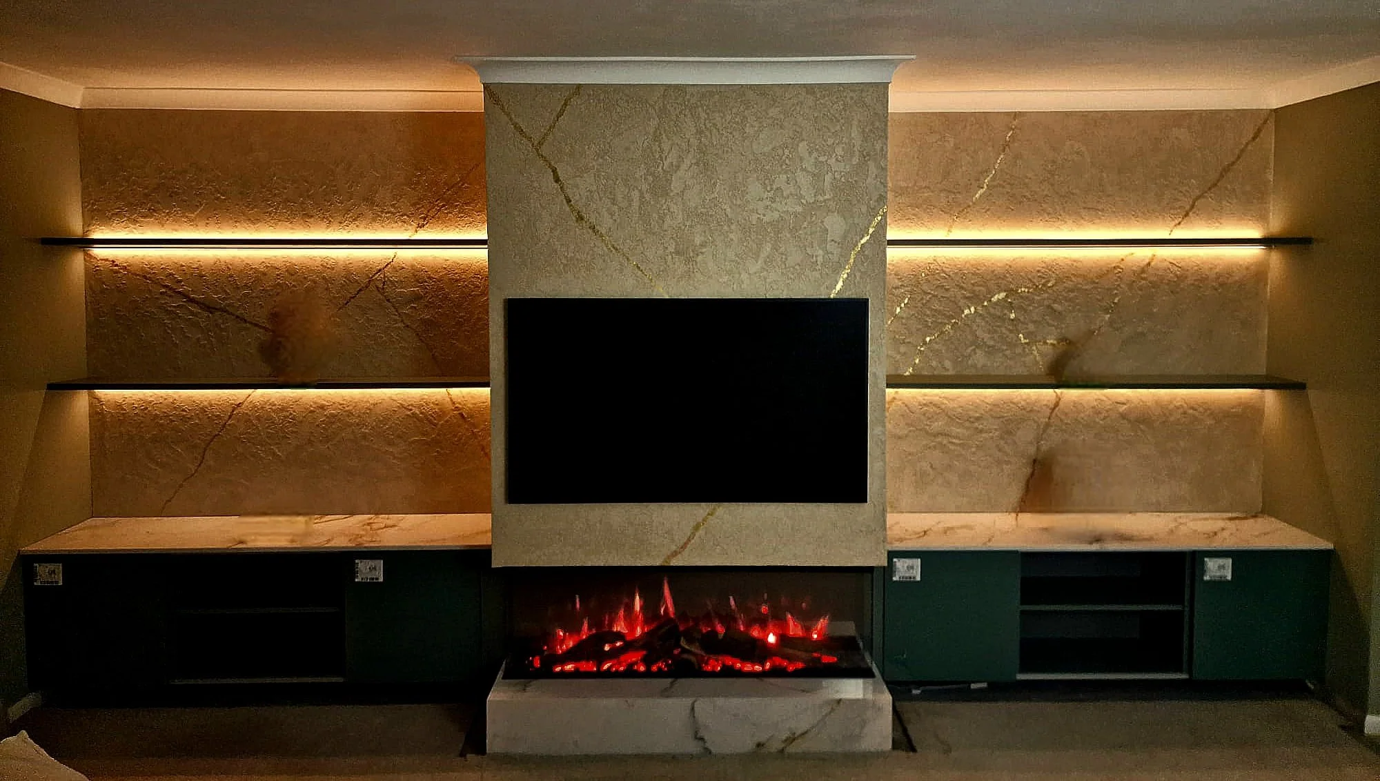 Modern living room with a large flat-screen TV mounted on a marble wall, illuminated shelving with warm lighting, and a built-in fireplace with red and orange flames and glowing logs beneath the TV.