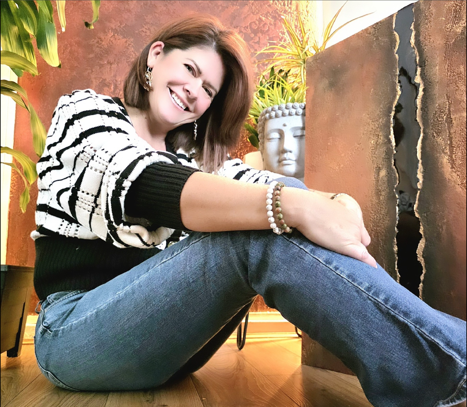 A smiling woman with shoulder-length brown hair, wearing a black and white striped sweater, blue jeans, and jewelry, sitting on the wooden floor in a cozy, decorated room with plants and a white sculpture of a face in the background.