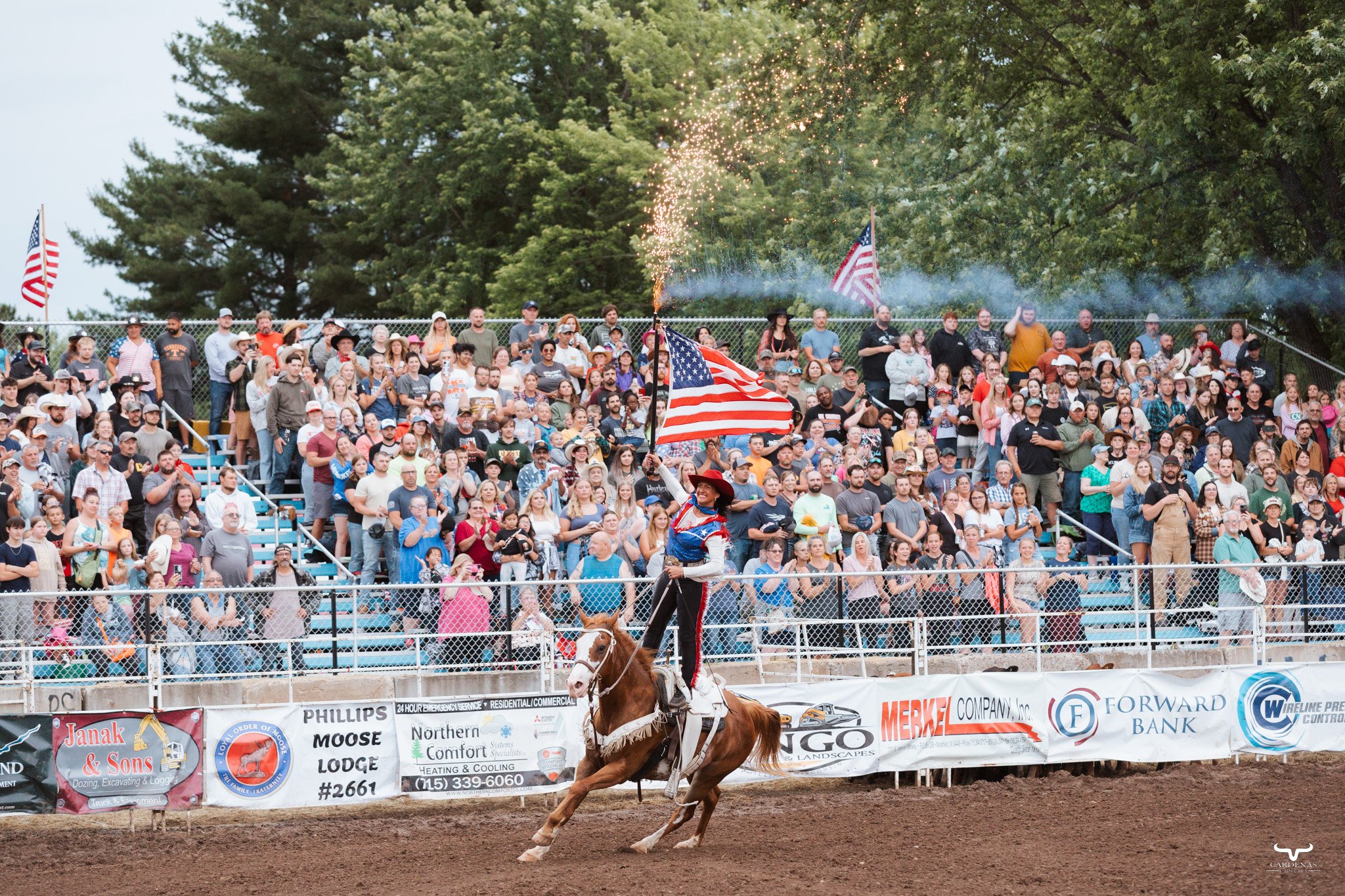 A rodeo event with a woman rider on a galloping horse holding a large American flag, in front of a crowd in the stands, with flags and banners along the fence.