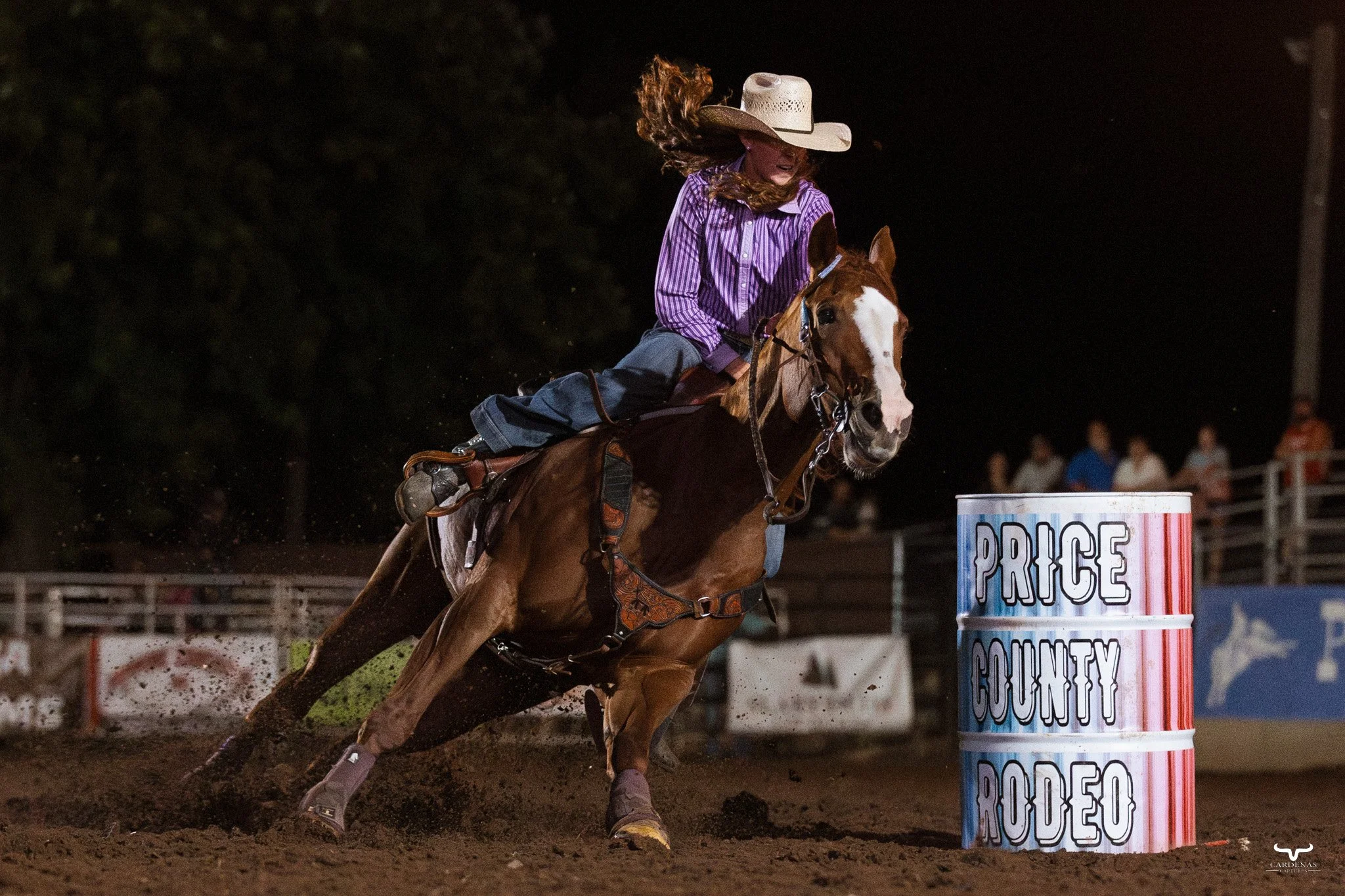 Cowgirl in purple striped shirt and cowboy hat riding a horse around a barrel labeled "Price County Rodeo" at a nighttime rodeo event.