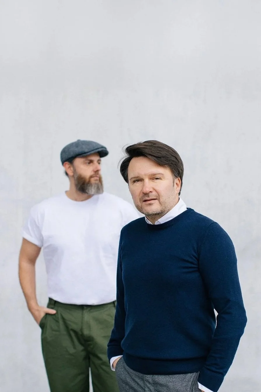 Zwei Männer stehen vor einer hellen Wand, einer in einem blauen Pullover und grauen Hosen, der andere in einem weißen T-Shirt, grünen Hosen und einer grauen Kappe.