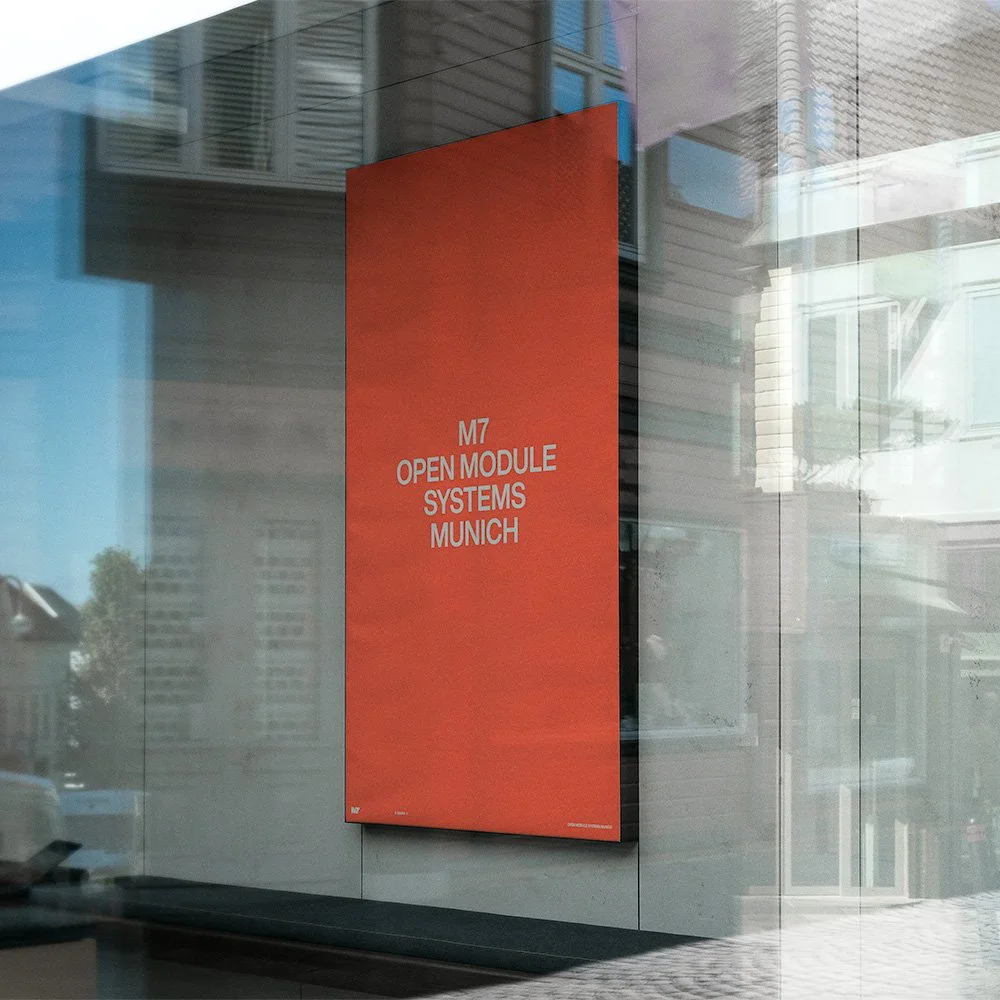 Ein rot-orangefarbenes Plakat in einer Glasvitrine mit weißen Texten, auf dem steht: "M7 OPEN MODULE SYSTEMS MUNICH". Die Umgebung zeigt reflektierende Fensterscheiben und eine Straße mit Gebäuden.