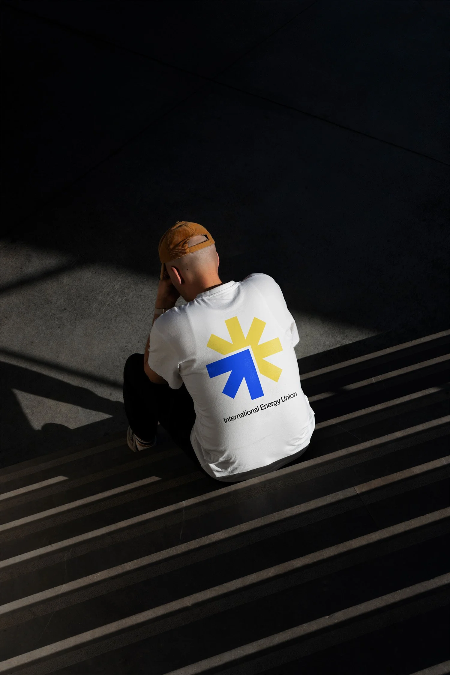 Junge Person sitzt auf Treppe, trägt ein weißes T-Shirt mit dem Logo der International Energy Union und eine braune Kappe, mit dem Rücken zur Kamera, in einem dunklen Raum.