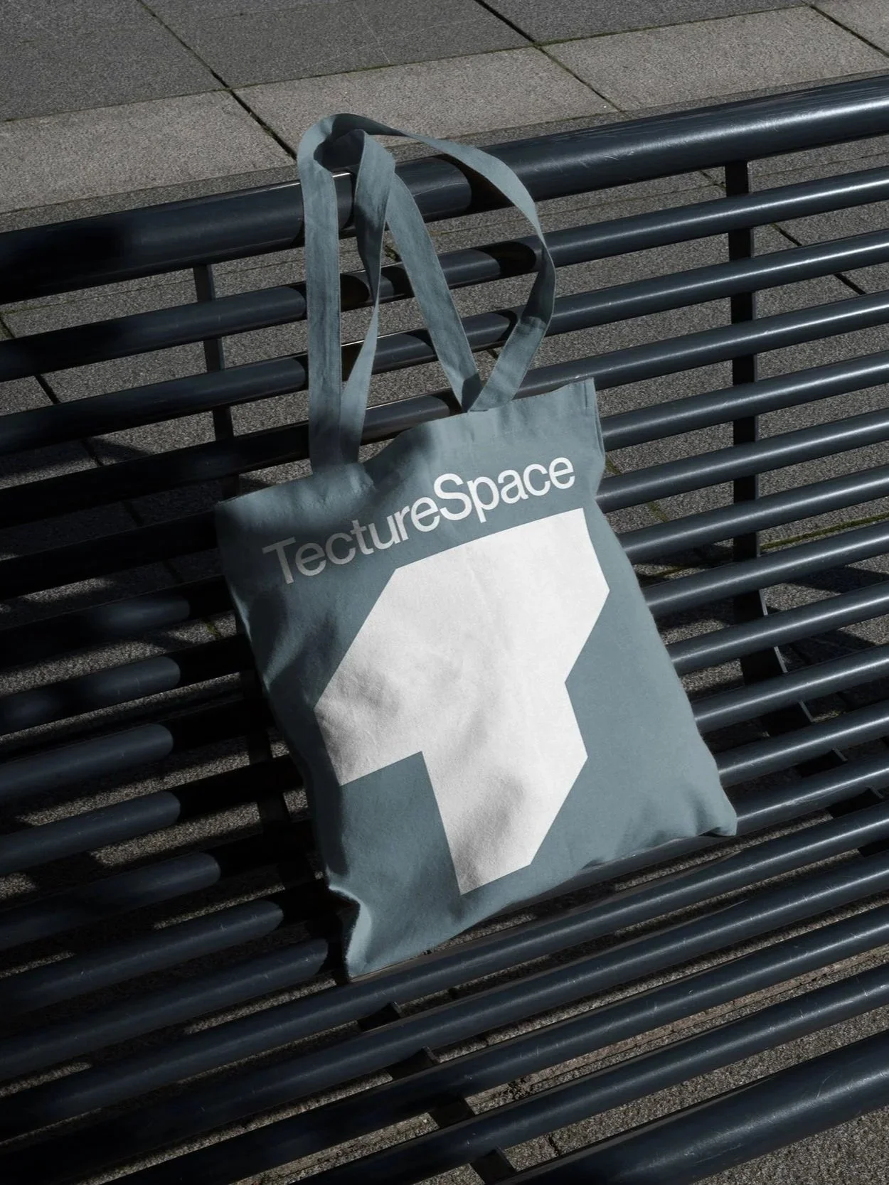 Eine graue Stofftasche mit dem Logo und Namen 'TectureSpace' liegt auf einer schwarzen Metallbank im Freien.