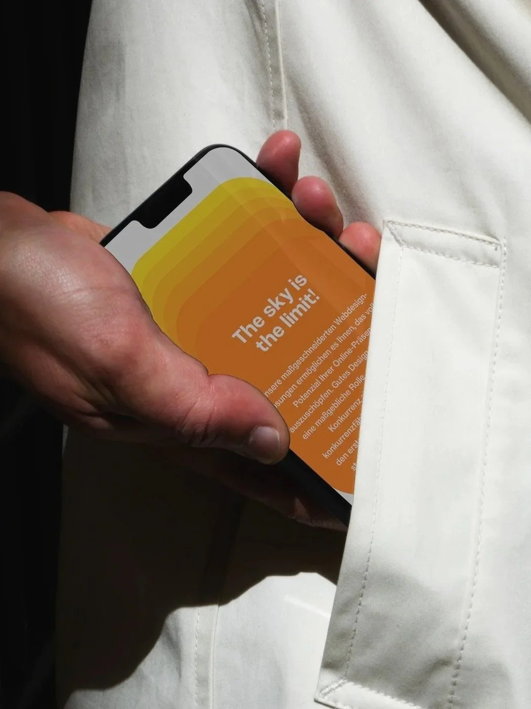Eine Hand hält ein Smartphone, das eine orangefarbene Seite mit weißen Texten zeigt. Im Hintergrund ein weißes Hemd. Auf dem Bildschirm steht: 'The sky is the limit!'