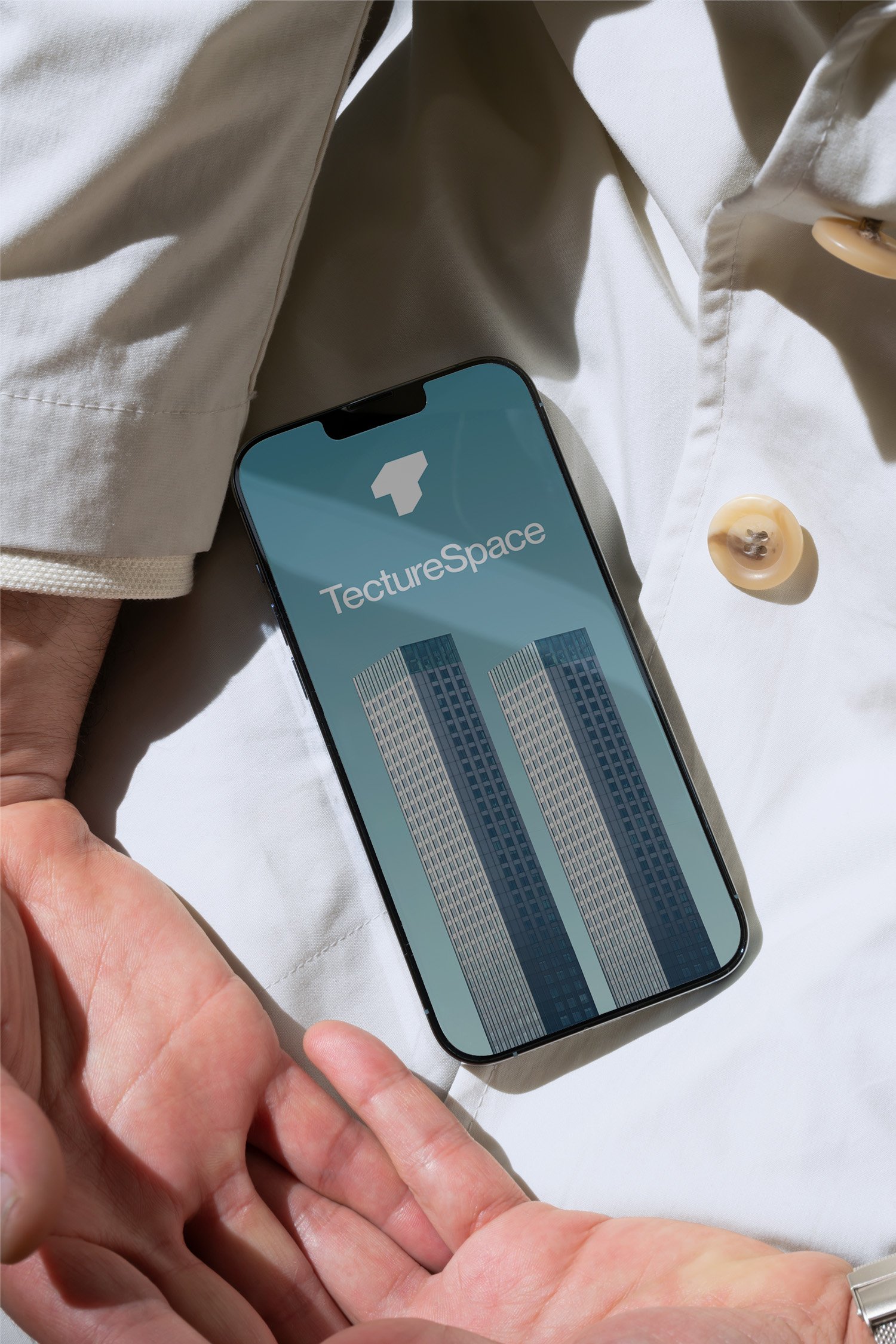 Nahaufnahme eines Smartphones mit der App 'TectureSpace' auf dem Bildschirm, gehalten von einer Person über einem weißen Stoffhintergrund, nahe einer weißen Jacke mit Knöpfen.