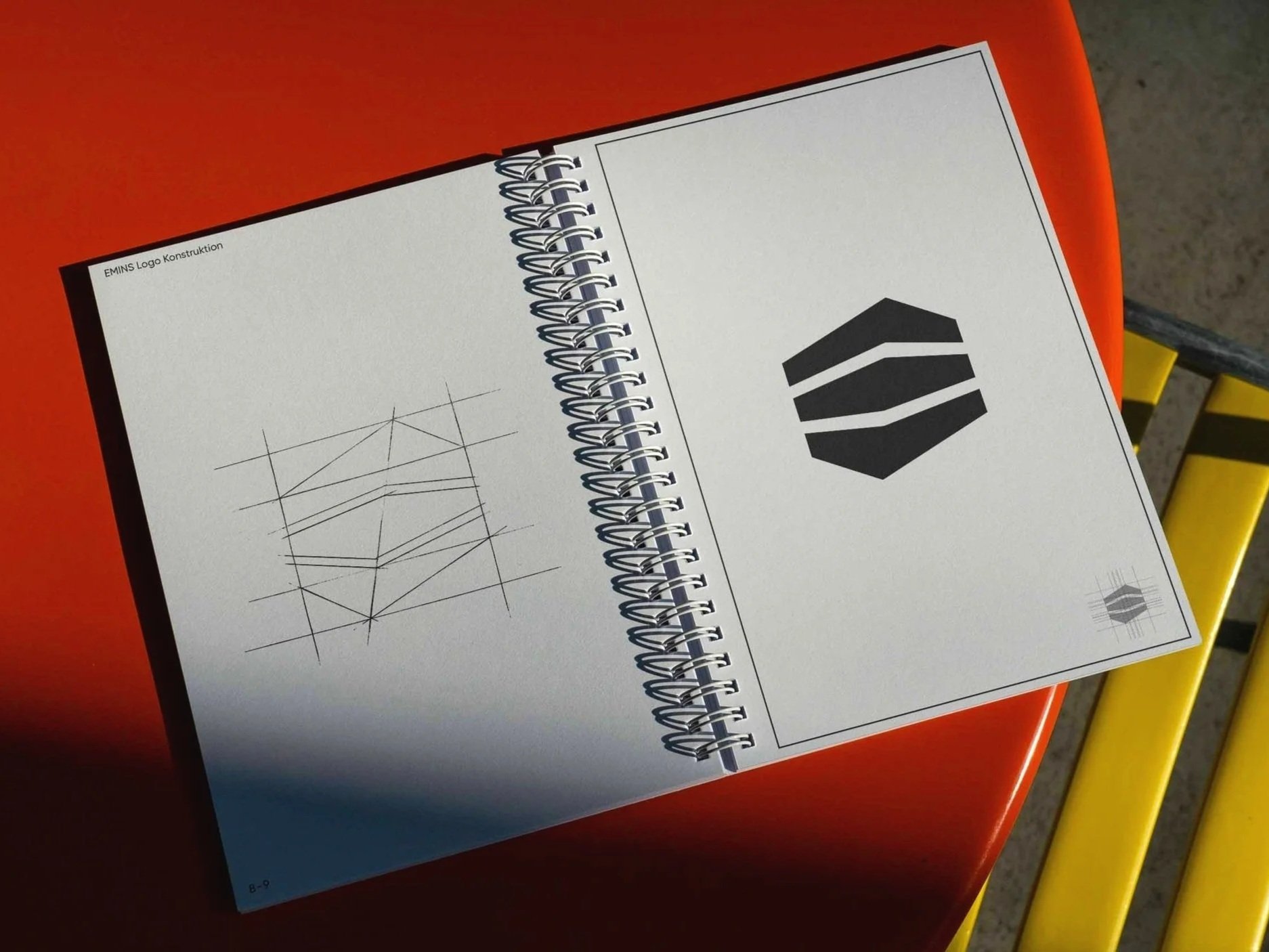Offenes spiralgebundenes Notizbuch mit technischen Zeichnungen und einem Logo auf der rechten Seite, gelegen auf einem roten Tisch. Der Hintergrund zeigt eine gelbe Sitzgelegenheit.