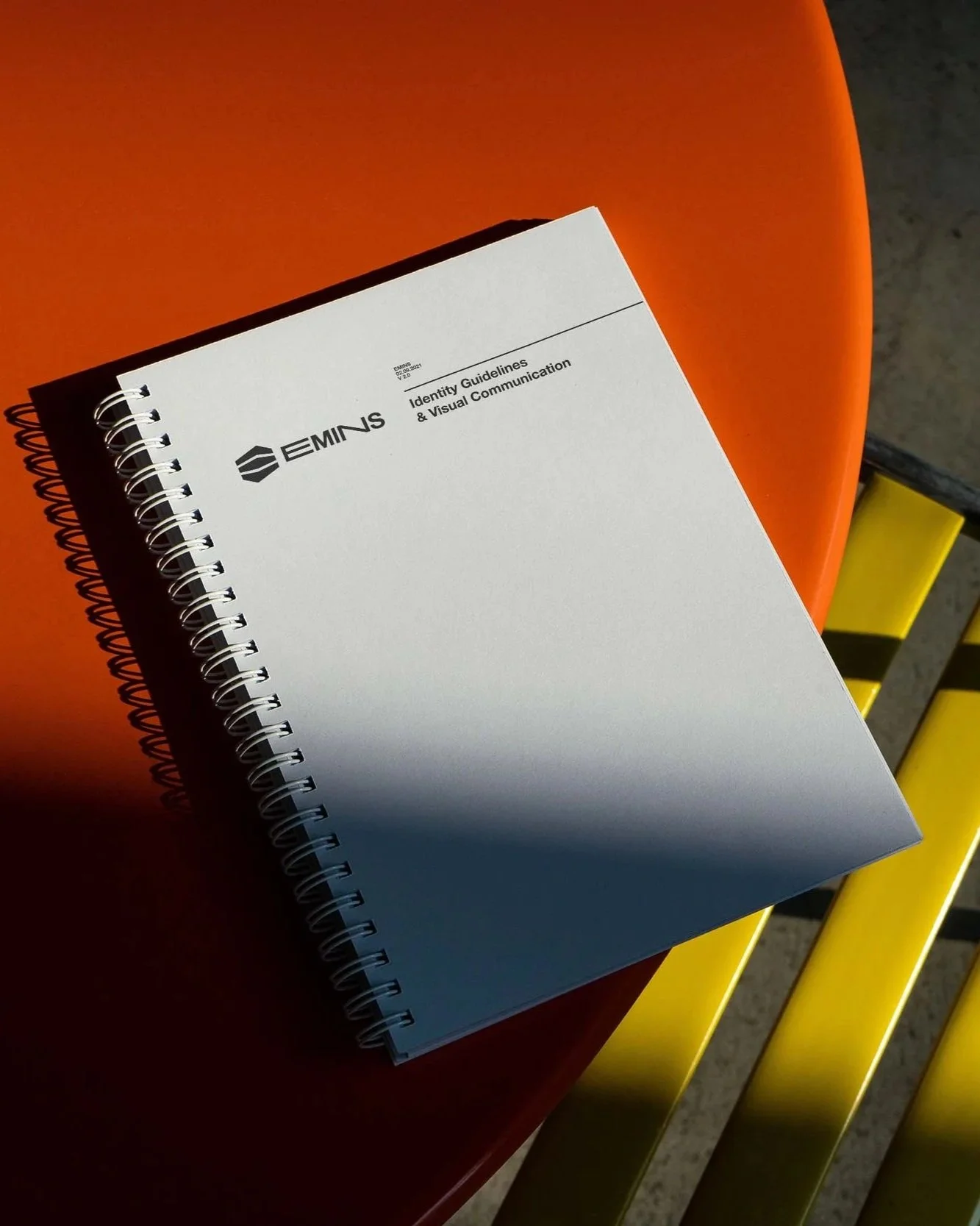 Ein Spiral gebundenes Whitepaper mit dem Titel 'EMINS' liegt auf einem orangefarbenen Tisch, der auf einem gelben Stuhl steht.