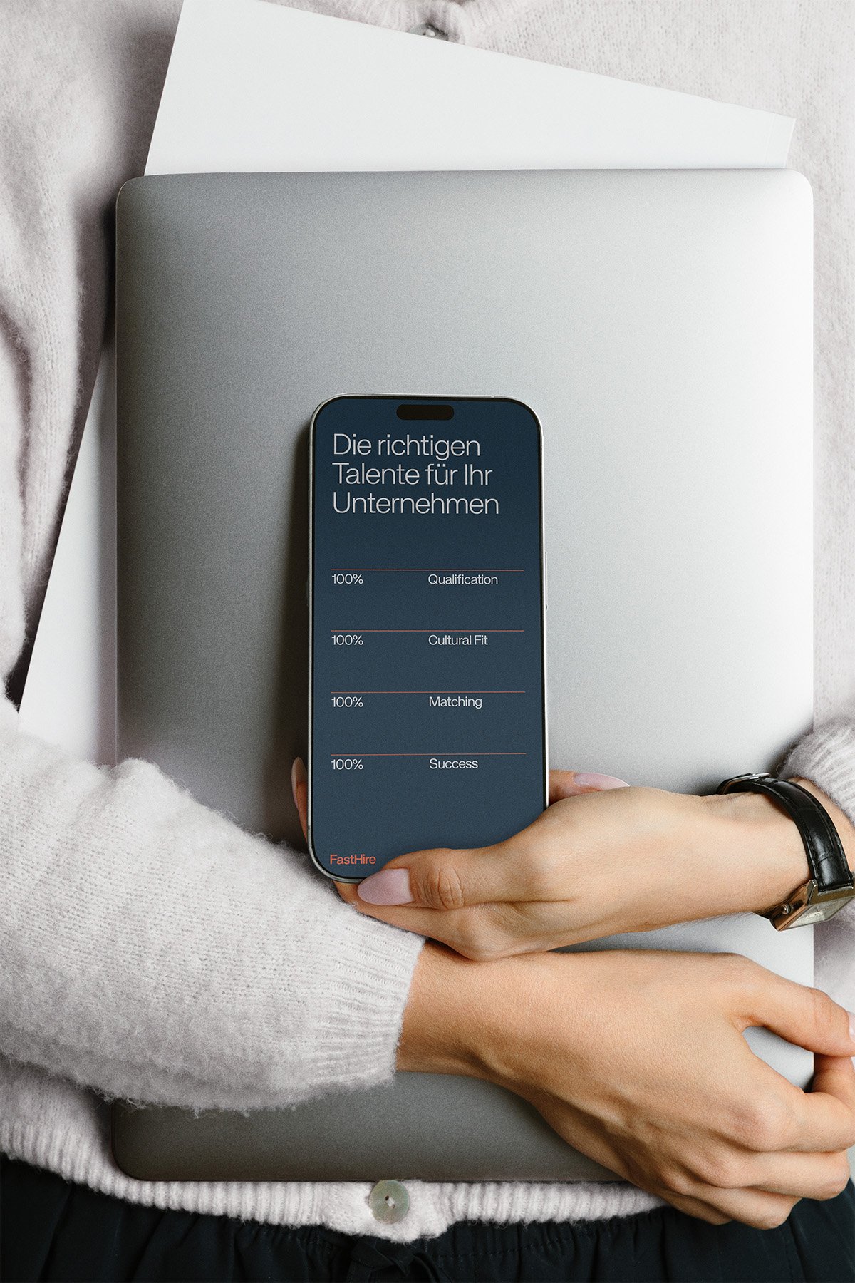 Eine Person hält ein Smartphone mit einer Anzeige, die 'Die richtigen Talente für Ihr Unternehmen' auf Deutsch zeigt, während sie auf einem Laptop liegt, auf dem ein Blatt Papier liegt, alles auf einem weißen Stoffbett.