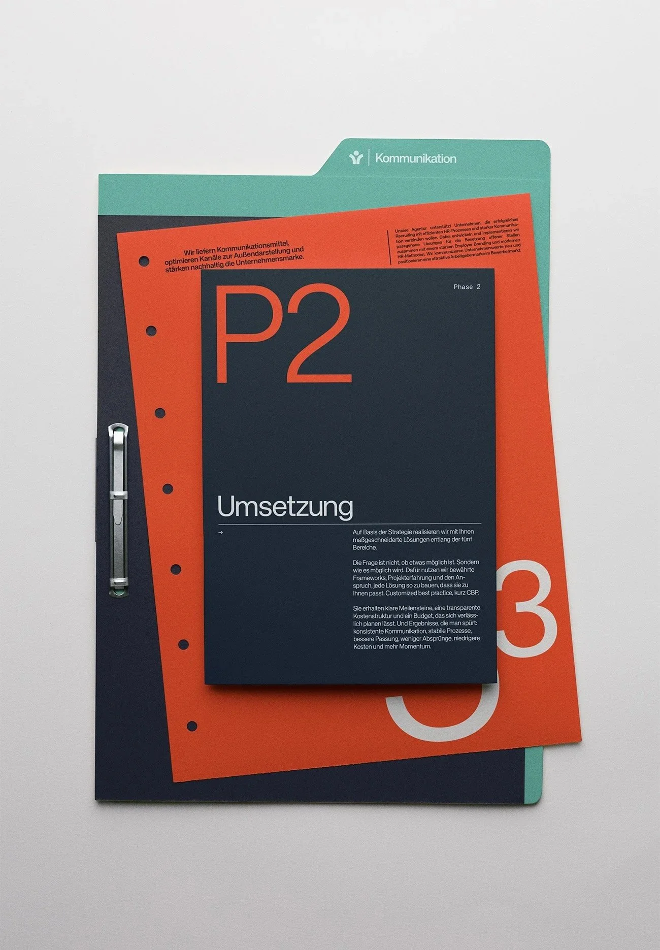 Drei farbige Dokumentenordner auf einem weißen Tisch, einer ist türkis, einer orange und einer dunkelblau. Auf dem dunkelblauen Ordner liegt ein schwarzes Heft mit der Aufschrift 'P2 Umsetzung'.