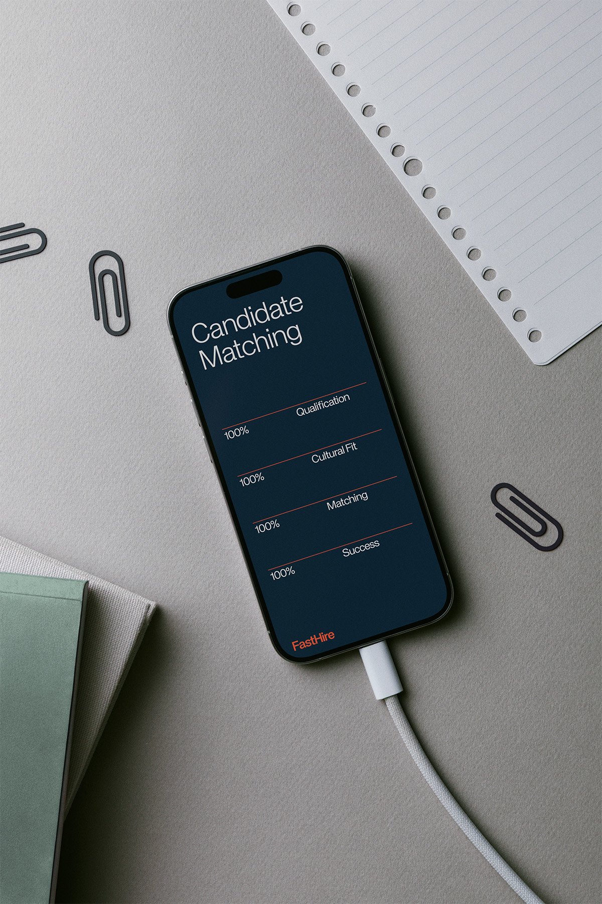 Smartphone auf einem Schreibtisch zeigt 'Candidate Matching' mit Vollständigkeit bei Qualifikation, kultureller Passung, Matching und Erfolg, verbunden mit einem Ladekabel. Es liegen Notizbücher, Papierklammern und ein Notizblock auf dem Tisch.