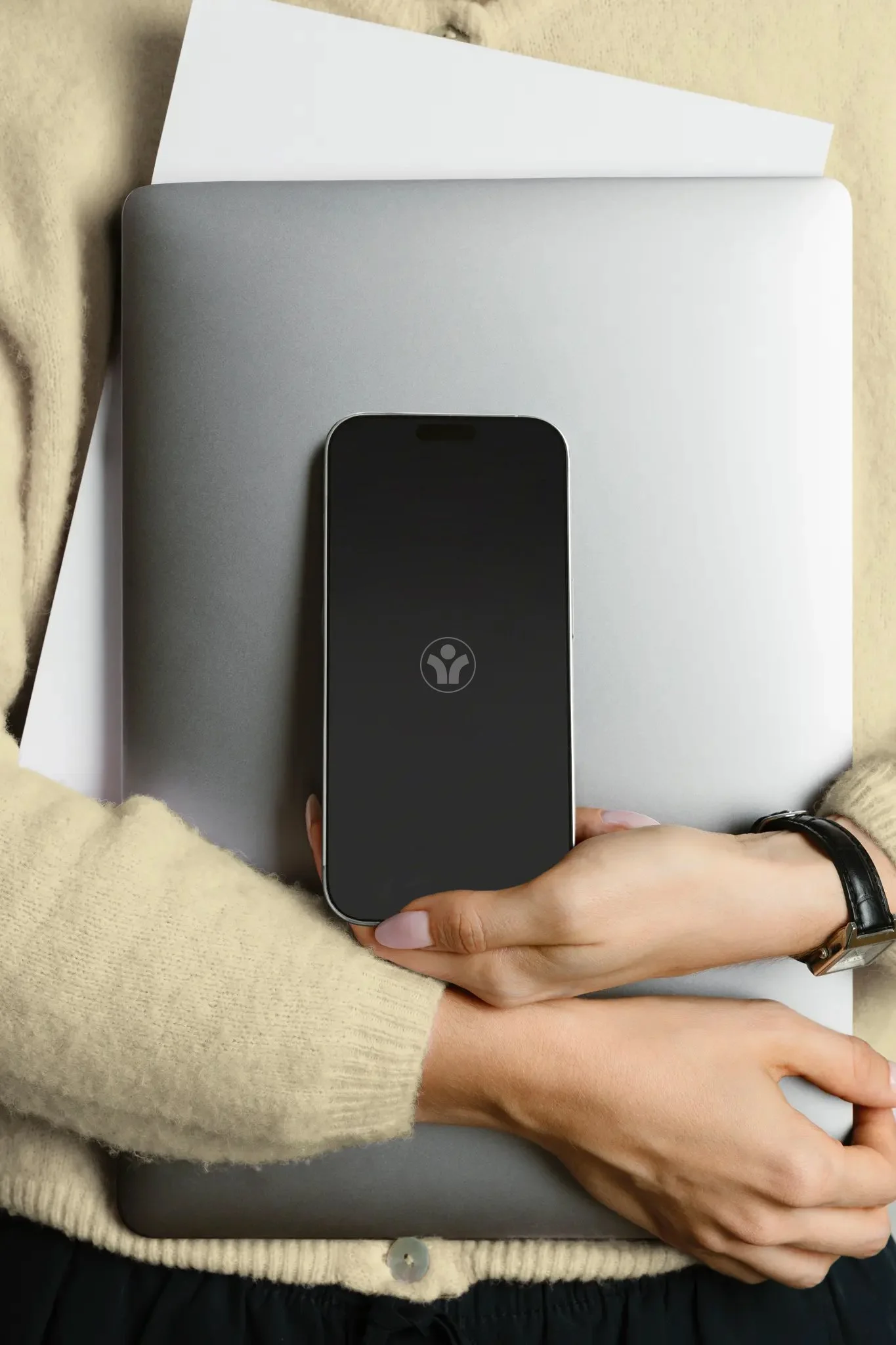 Nahaufnahme einer Person, die ein Smartphone auf einem Laptop trägt, auf einem Tisch liegt, umgeben von einer beige Strickjacke und einem schwarzen Uhrenarmband.