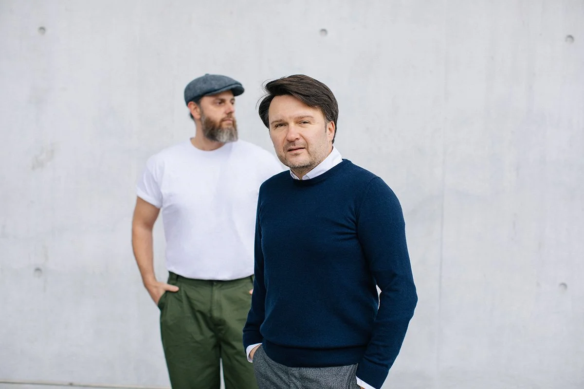 Zwei Männer stehen vor einer hellen Betonwand. Der Mann im Vordergrund trägt einen dunklen Pullover und sieht direkt in die Kamera, während der Mann im Hintergrund einen Hut und ein weißes T-Shirt trägt und zur Seite schaut.