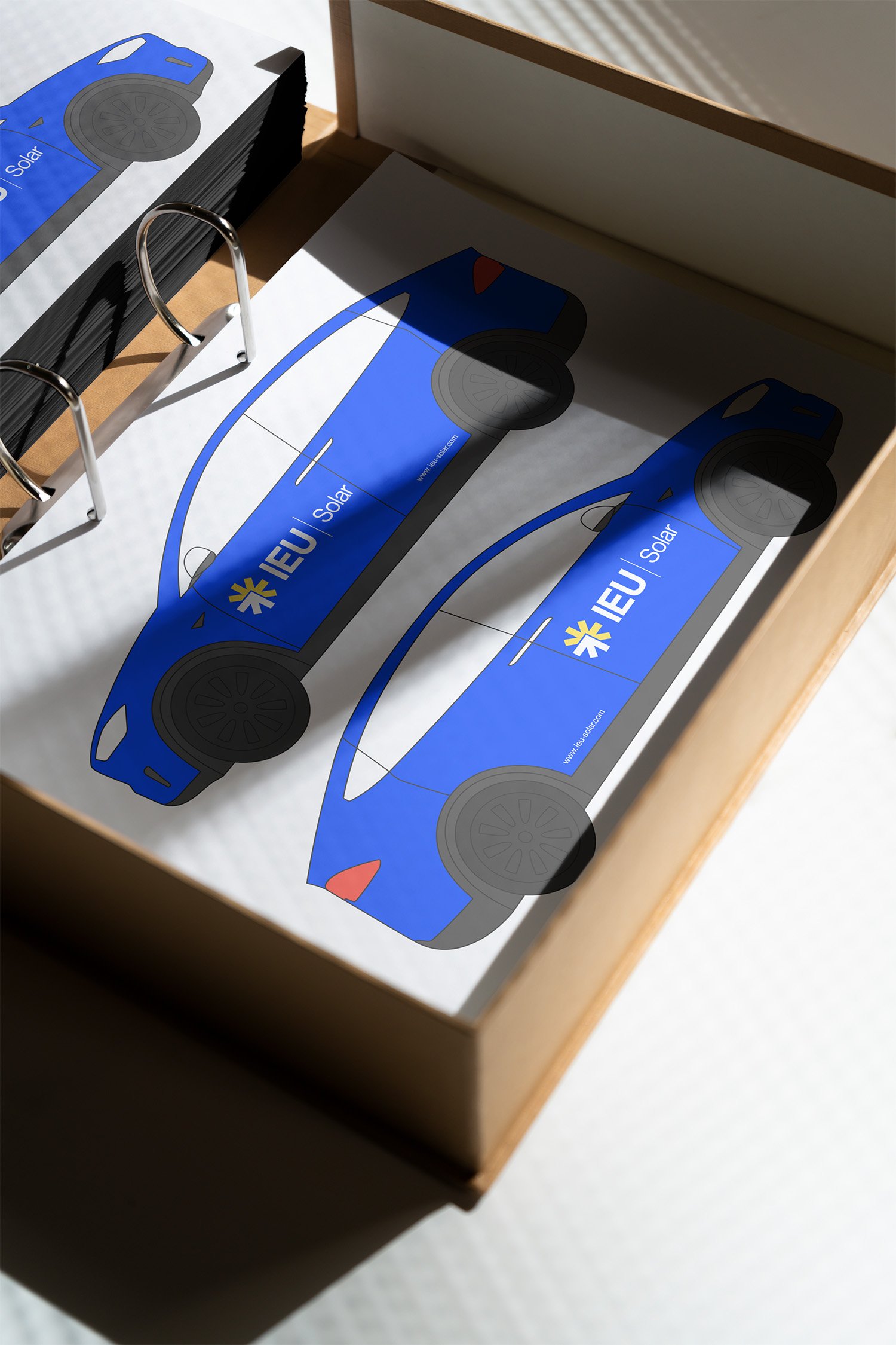 Ein offenes Pappkarton mit gedruckten blauen Solarwagen-Designs. Darunter befindet sich eine weiße Papier-ähnliche Unterlage mit den gleichen Designs, die auf einem Klemmbrett befestigt ist.