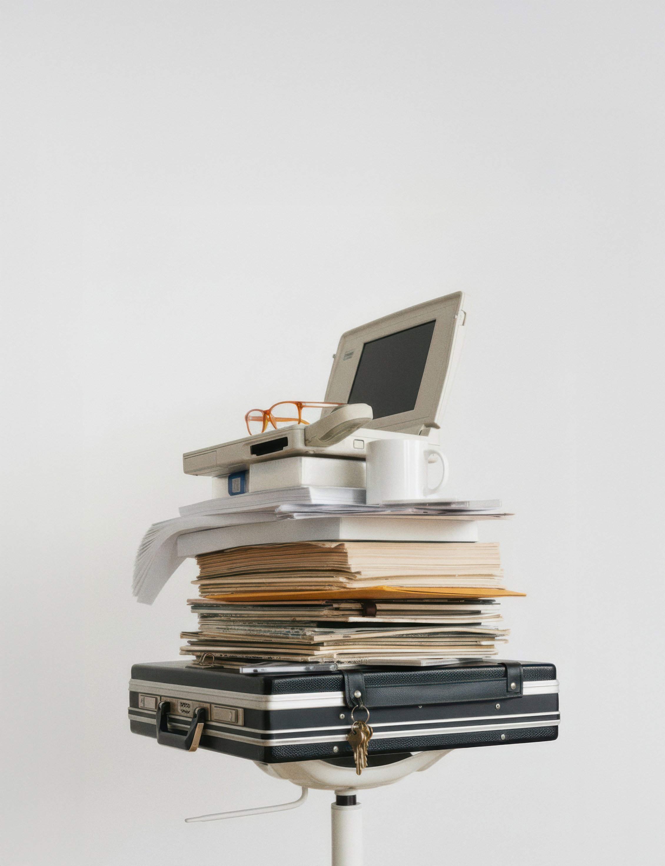 Ein aufgeräumter Schreibtischstuhl mit mehreren gestapelten Akten, einem alten Computer und einer Tasse, dazu eine Brille und ein Schlüssel hängen am Koffer.