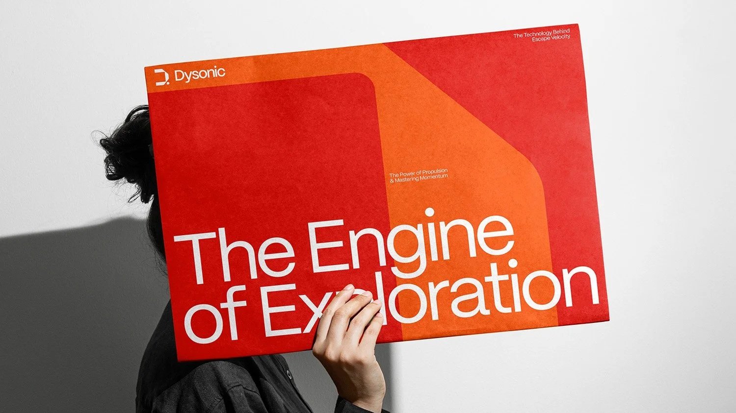 Eine Person hält ein großes rotes Plakat mit dem Titel 'The Engine of Exploration', teilweise den Körper verdeckend, vor einem weißen Hintergrund.