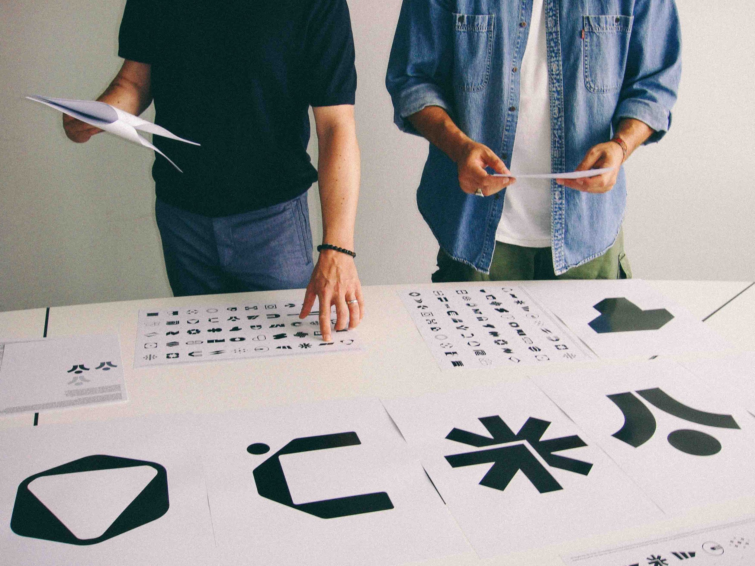 Zwei Personen stehen über einem weißen Tisch mit großen Buchstaben und Symbolen. Die eine Person zeigt auf einen Bereich mit schwarzem Text auf weißen Papierbögen. Die andere Person hält eine Papierlage in der Hand. Im Hintergrund eine graue Wand.