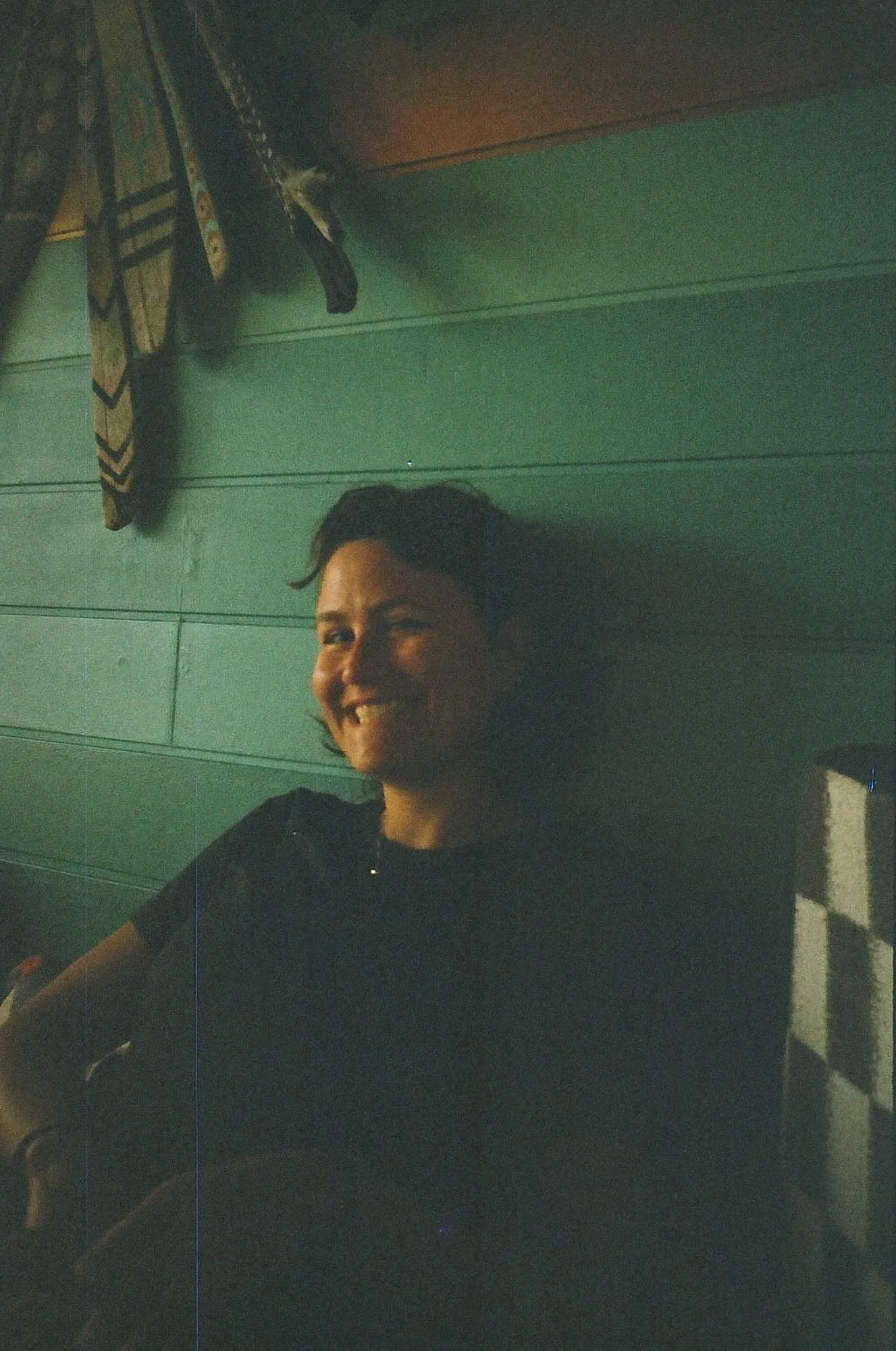 Portrait de Marie-Louise Chappuis, souriante dans une pièce sombre, assise contre un mur vert, avec des vêtements sombres et des éléments décoratifs en arrière-plan.