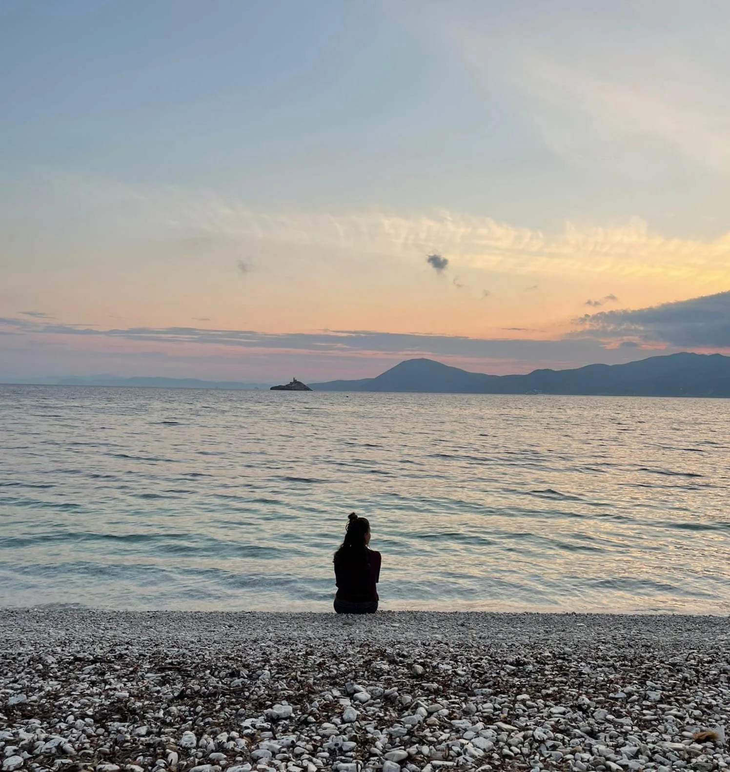 Personne assise sur une plage de galets, regardant la mer avec un coucher de soleil, avec des montagnes en arrière-plan.