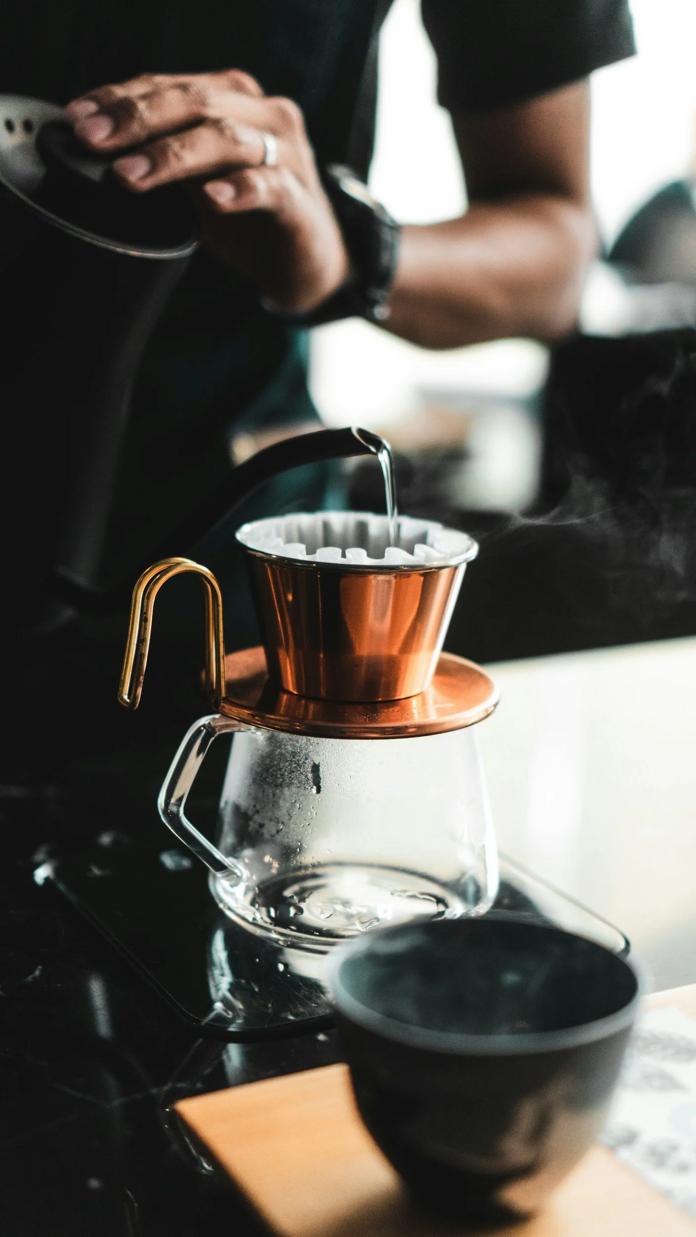 A person is brewing coffee using a pour-over method, with hot water being poured from a kettle into a copper-colored coffee dripper placed on top of a glass carafe. A black bowl is in the foreground on a wooden surface.