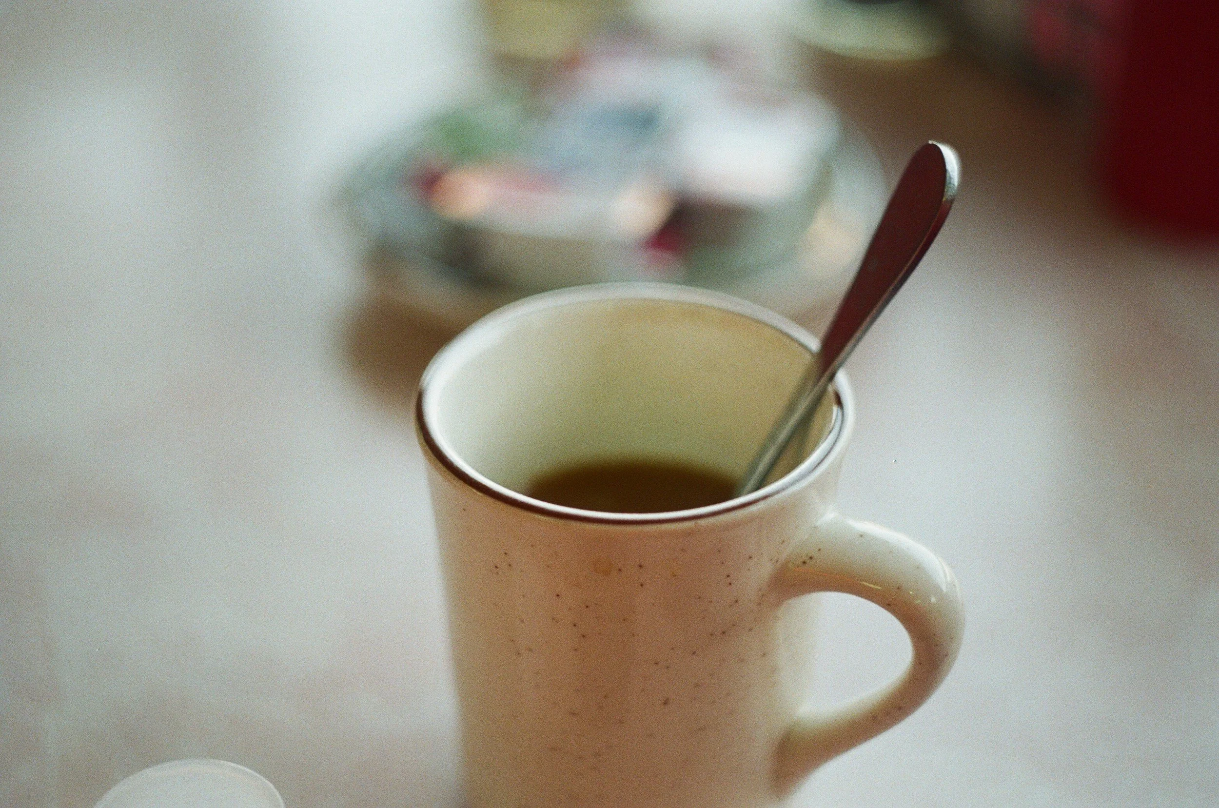 Close-up of an off-white speckled ceramic mug filled with a hot beverage, with a metal spoon inside, on a light surface with a blurred bowl of snacks or candy in the background.