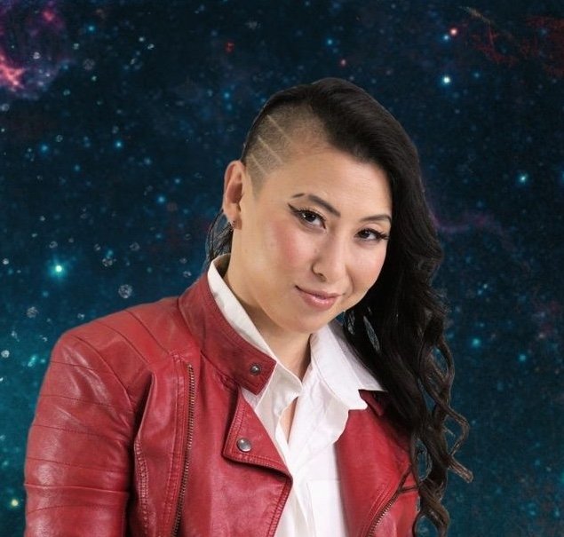 Erika Ishii, an Asian non-binary person with long black hair, their head shaved on one side, and a white shirt and red leather jacket, smiles in front of a dark blue starry sky.