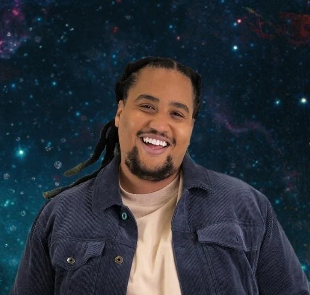 Lou Wilson, a Black man with black dreadlocks pulled back and beard on his chin, wearing a tan shirt and dark blue jacket, smiles in front of a dark blue starry sky.