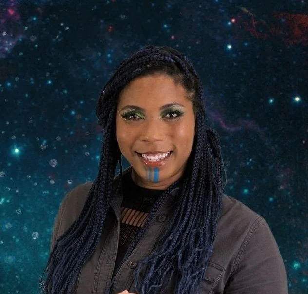 Aabria Iyengar, a Black woman with black and blue braids, a grey jacket, and blue makeup on her chin, smiles in front of a dark blue starry sky.