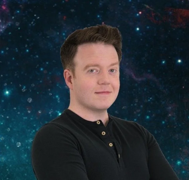 Brennan Lee Mulligan, a White man with dark red-brown hair and a black shirt with pale buttons smiles in front of a dark blue starry sky.