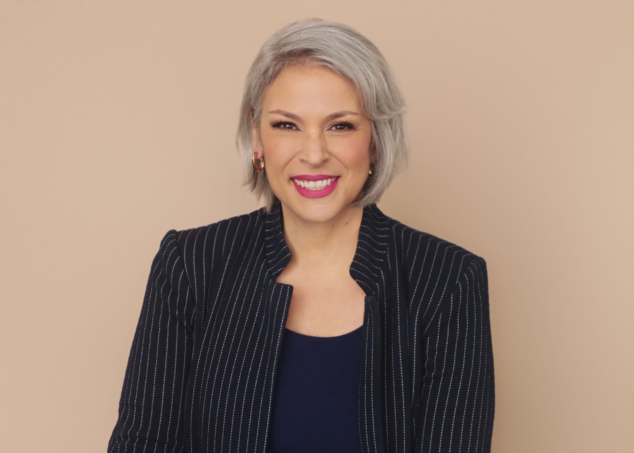 A woman with short gray hair smiling, wearing a black pinstripe blazer and gold earrings, against a beige background.