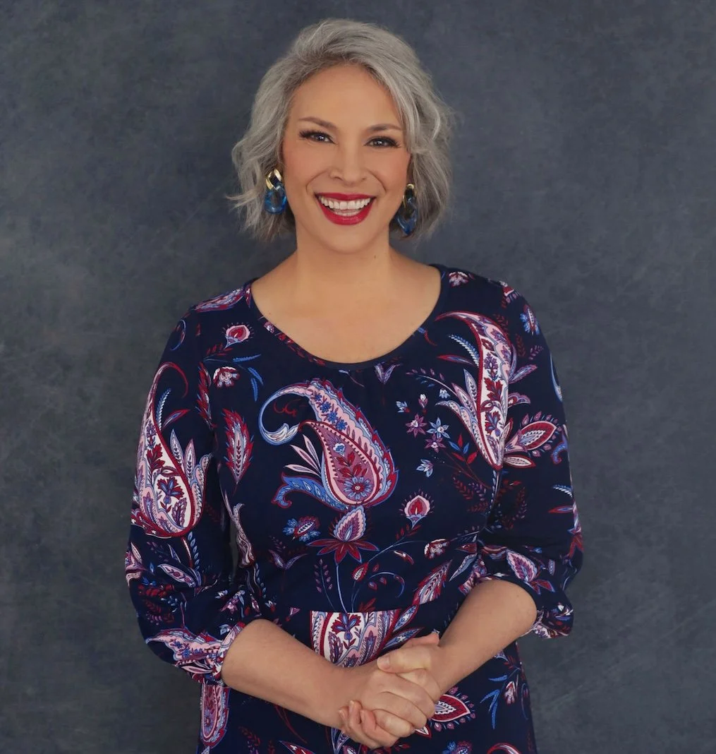 A woman with short gray hair smiling, wearing a dark floral dress with long sleeves and colorful earrings, standing against a dark textured background.